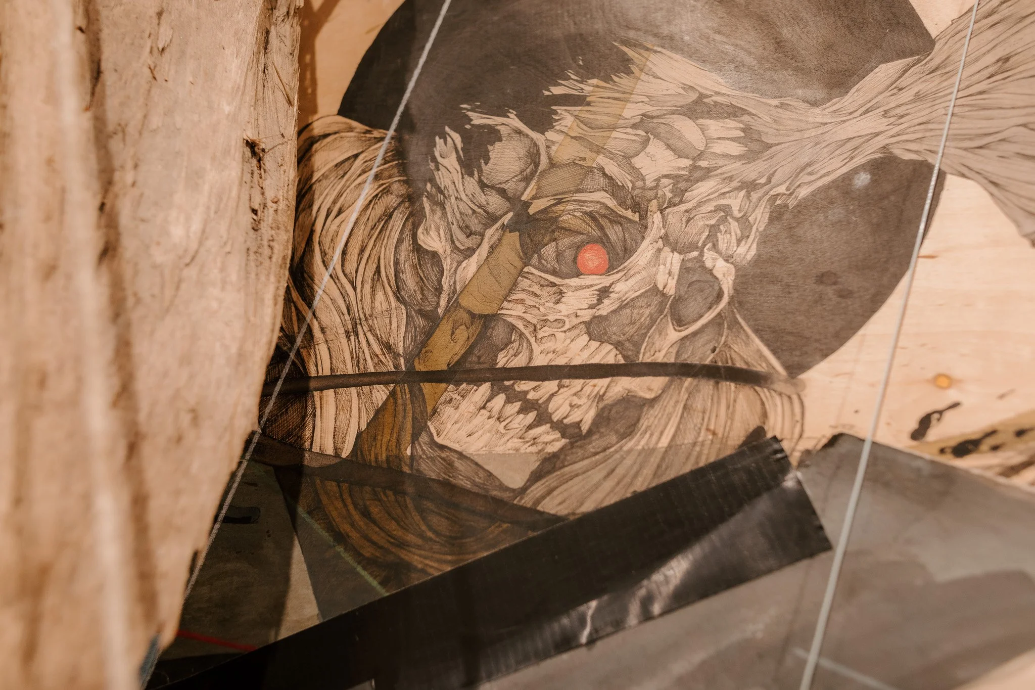 An art piece depicting a skull intertwined with branches and trees, with a small red circle near the eye socket, mounted on a wooden surface and protected by a glass cover.