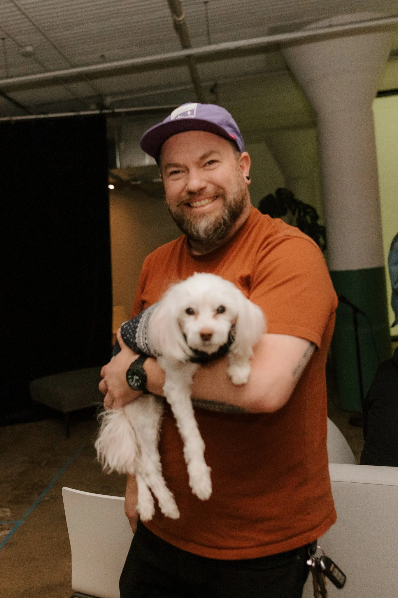 A smiling man with a beard and tattoos wearing a purple cap, holding a small white dog with floppy ears and a black sweater in an indoor setting.