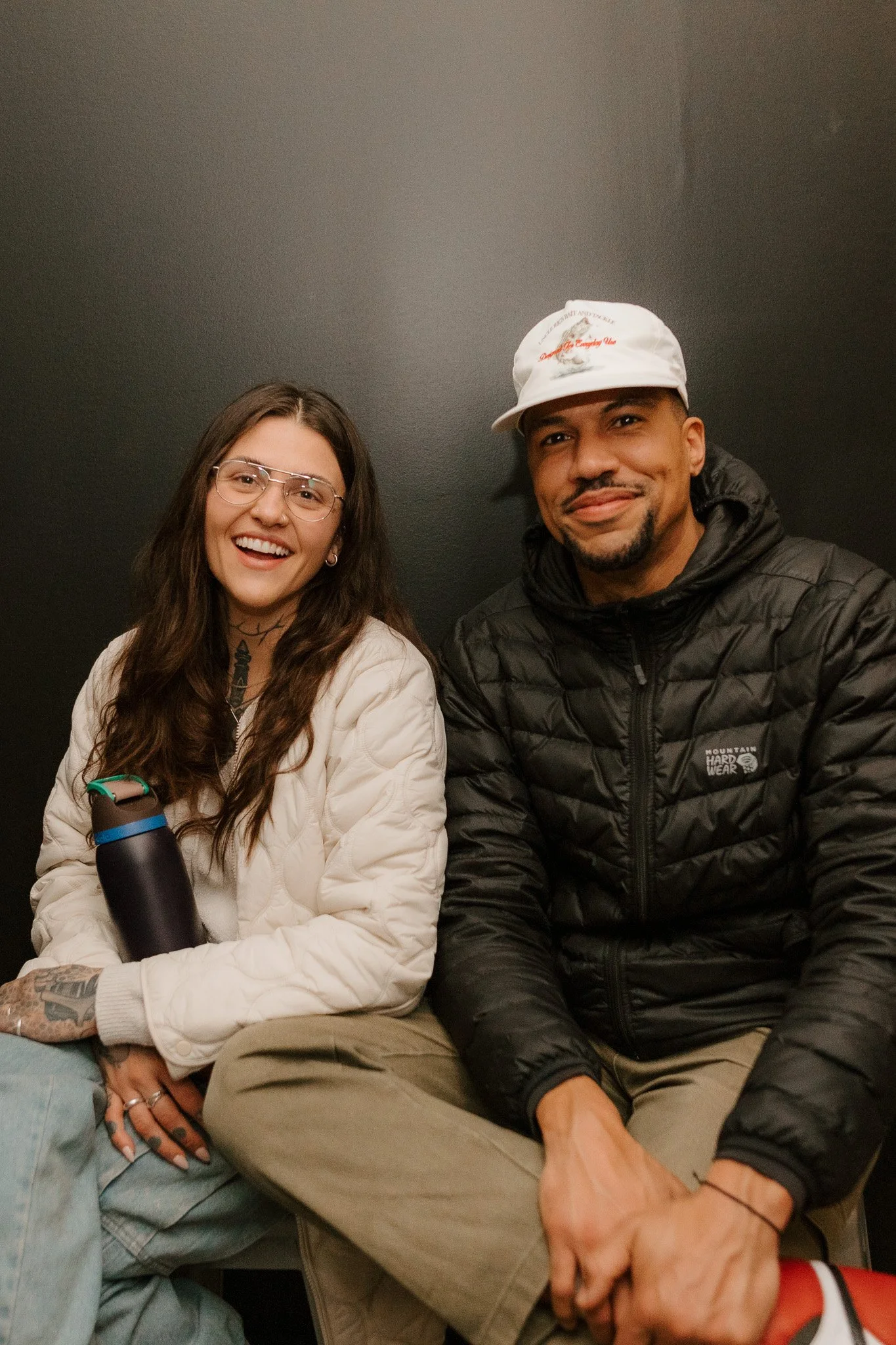 A smiling woman with long brown hair, wearing glasses, a white quilted jacket, and holding a water bottle. Beside her, a smiling man with a beard, wearing a white cap, black puffer jacket, and beige pants. They are sitting close together against a da