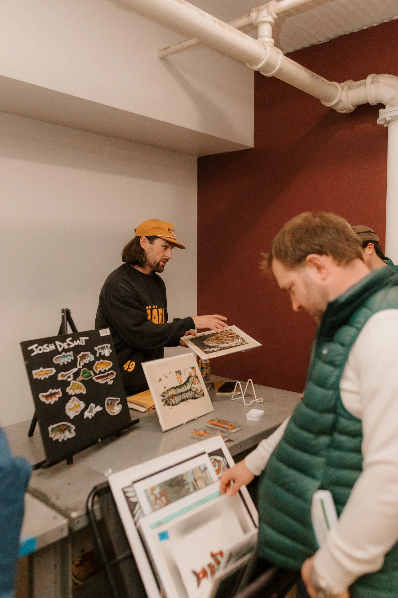 A man with long dark hair and a beard, wearing a black sweatshirt and yellow cap, shows artwork to a person at an event table. The table has stickers, illustrations, and a sign with the name 'Josh Desmit'.