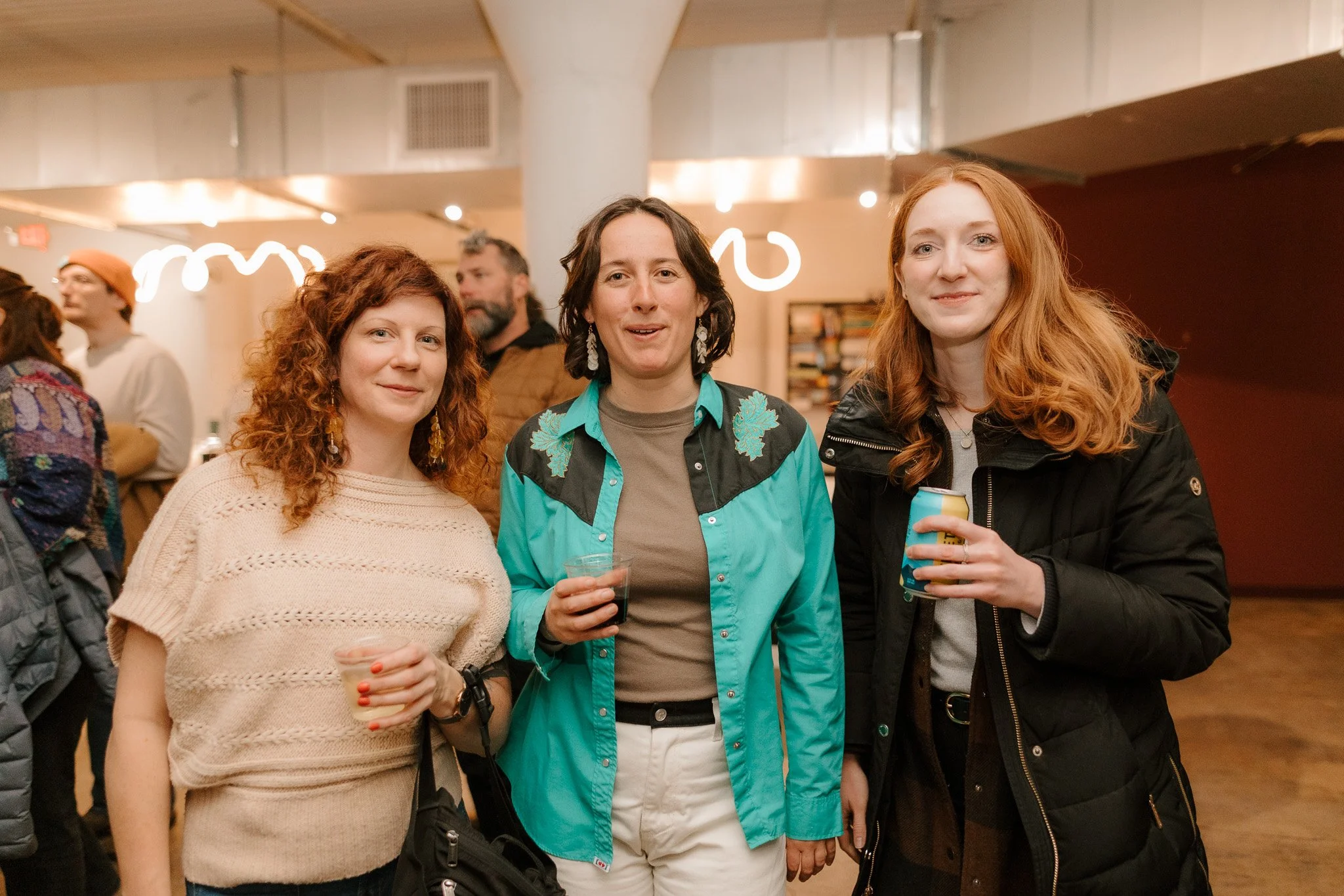 Three women with curly and wavy red hair holding drinks, standing together and smiling at an indoor social event with people in the background.