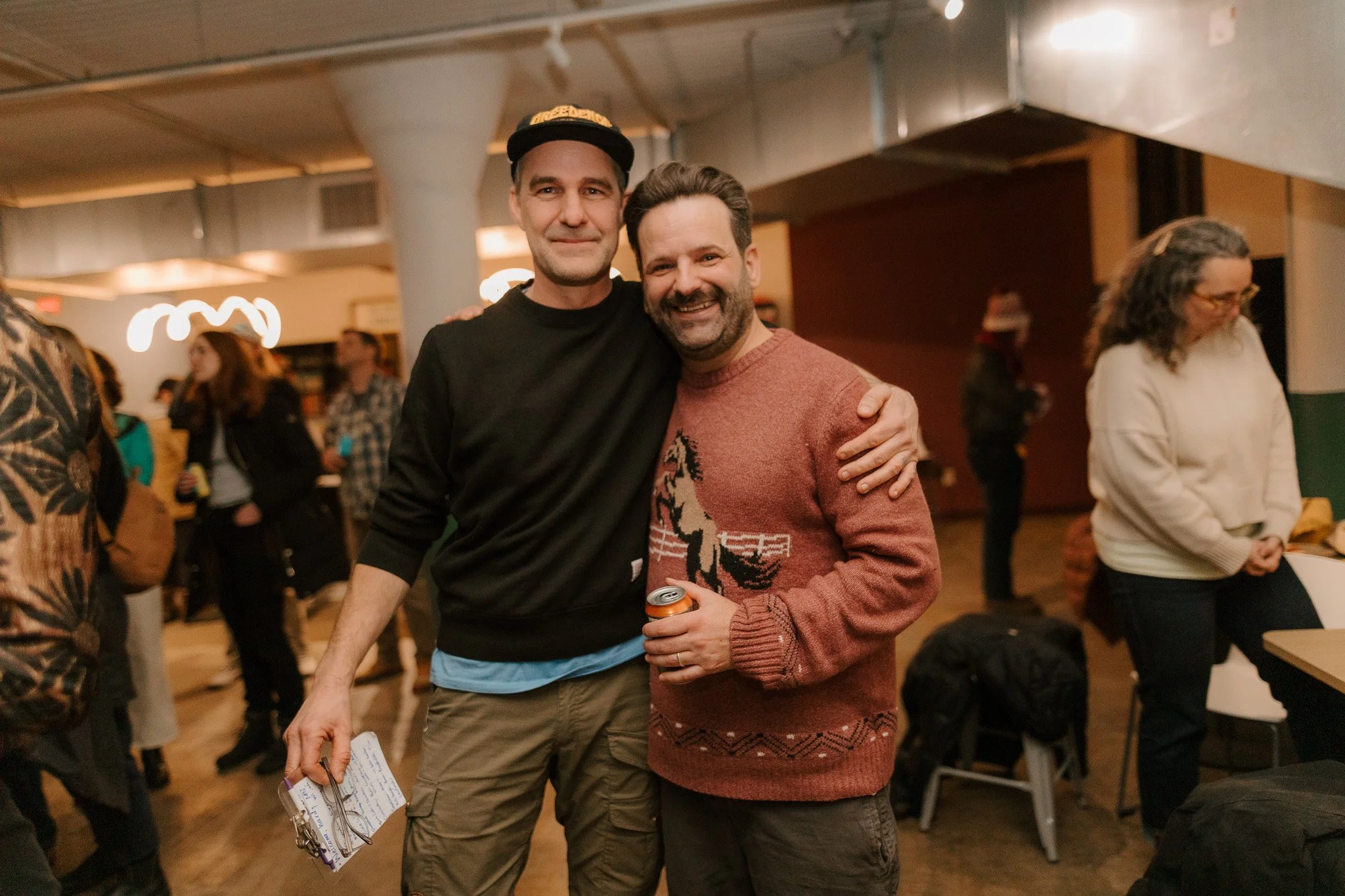 Two men smiling and embracing at a social gathering, one wearing a black sweatshirt with a hat, the other in a Christmas sweater with a horse design, holding a beverage in a can.