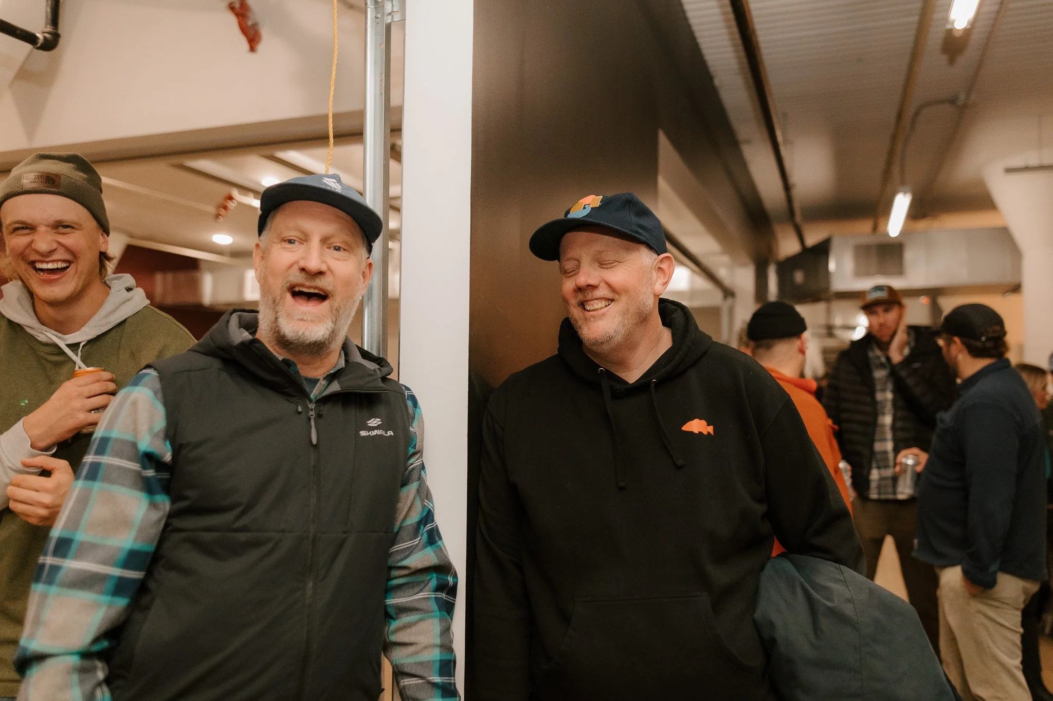 A group of men laughing and talking in a social indoor setting, some wearing black caps and jackets, with others in the background engaged in conversation.