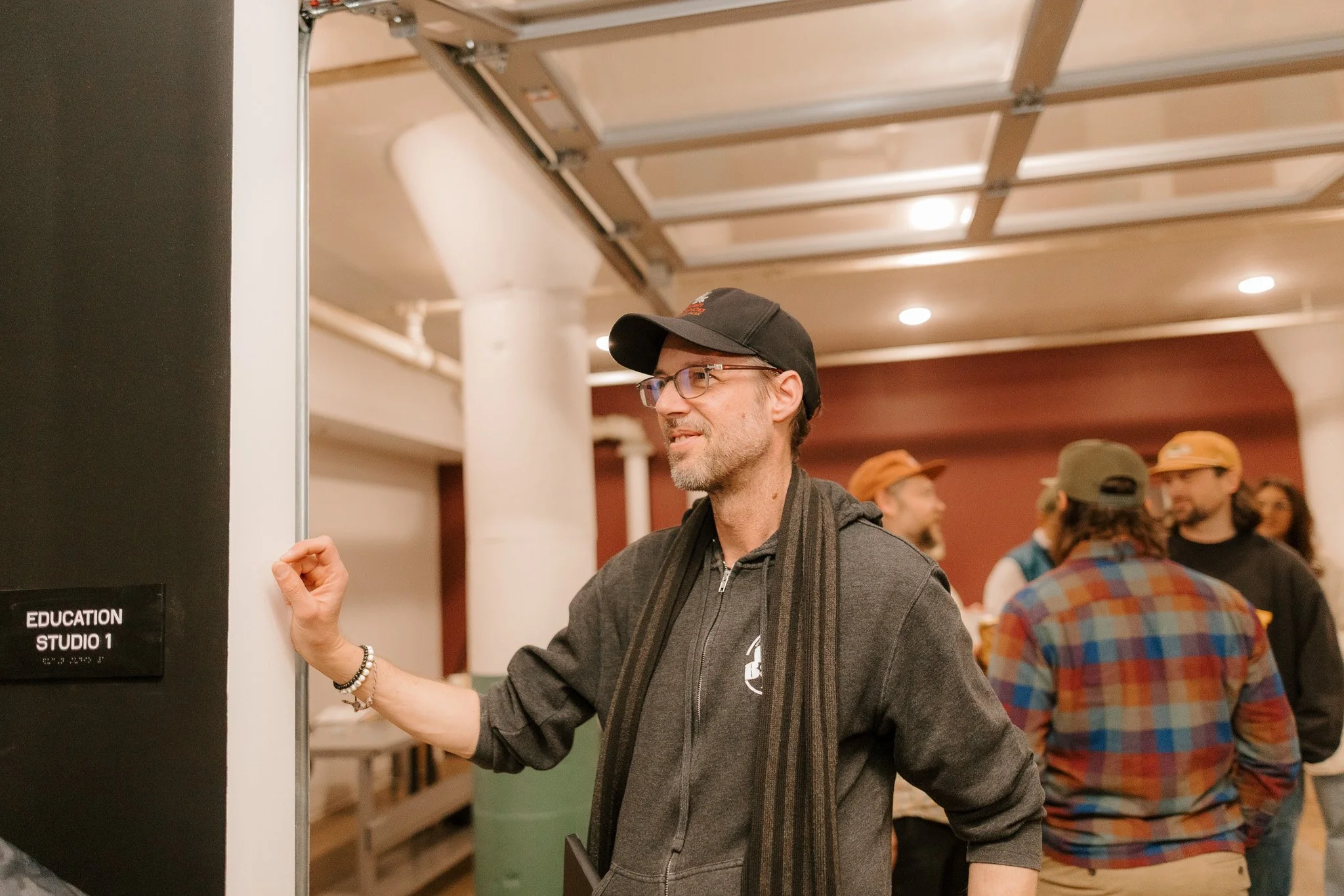 A man wearing glasses, a black cap, a gray hoodie, and a striped scarf stands with his arm resting on a door frame labeled 'Education Studio 1' while looking to the left. In the background, several people are conversing in a room with white columns a