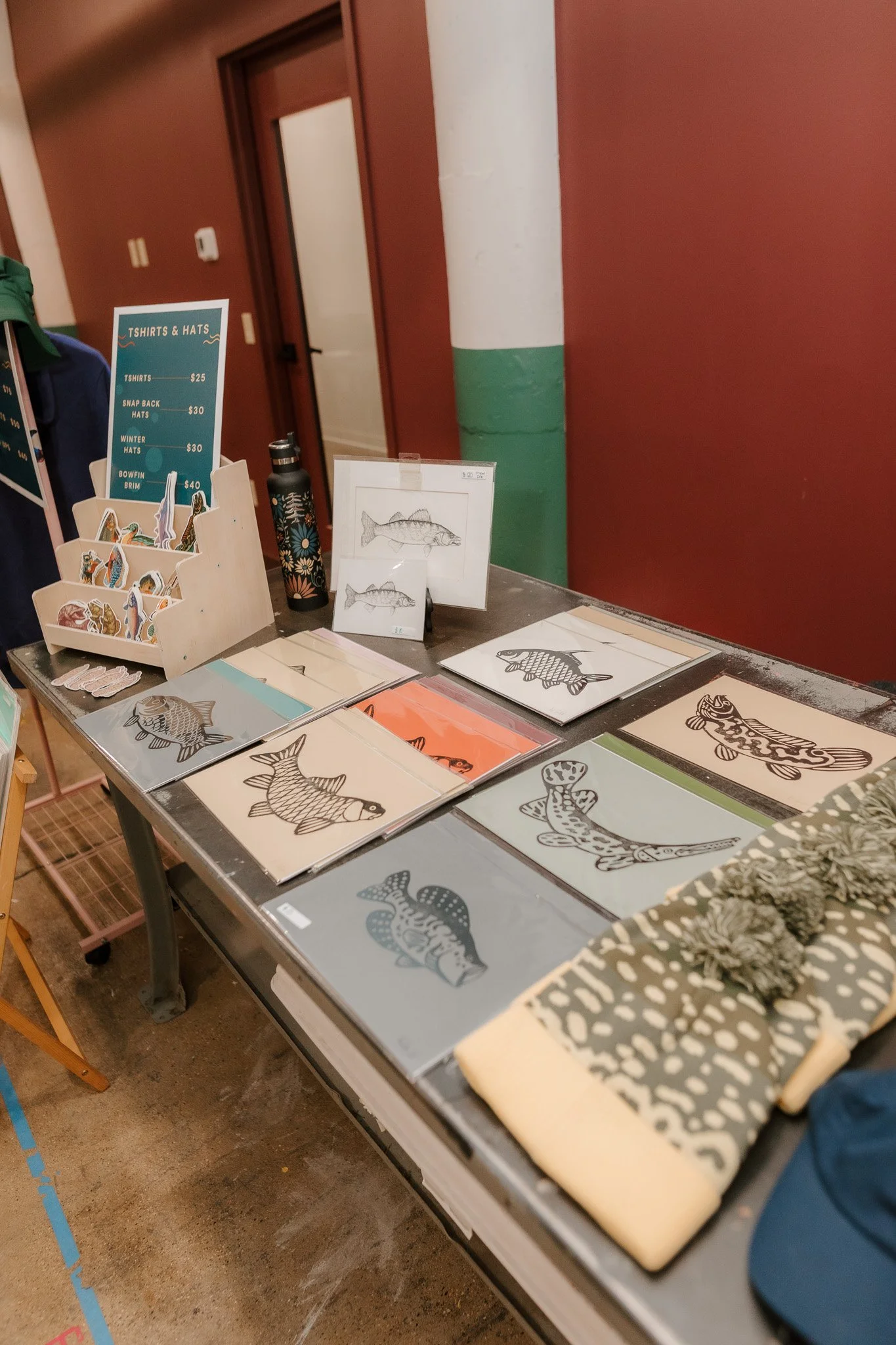 Table displaying fish-themed artwork, stickers, and hats, with a sign listing T-shirt and hat prices, set against a red wall with a green and white painted column.
