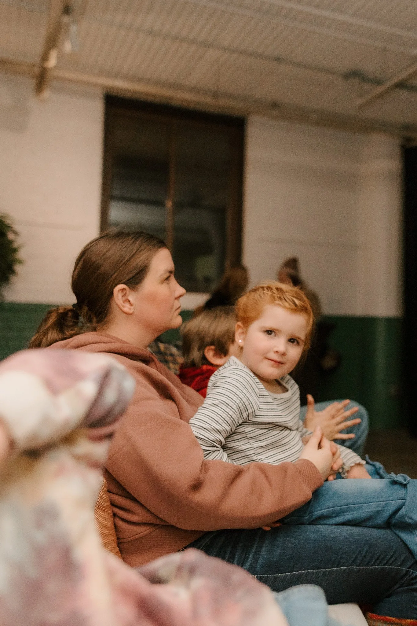 A woman and young girl sitting indoors with other children, watching something off-camera. The girl has red hair and is wearing a striped shirt, looking at the camera with a slight smile. The woman has brown hair pulled back and is wearing a brown ho