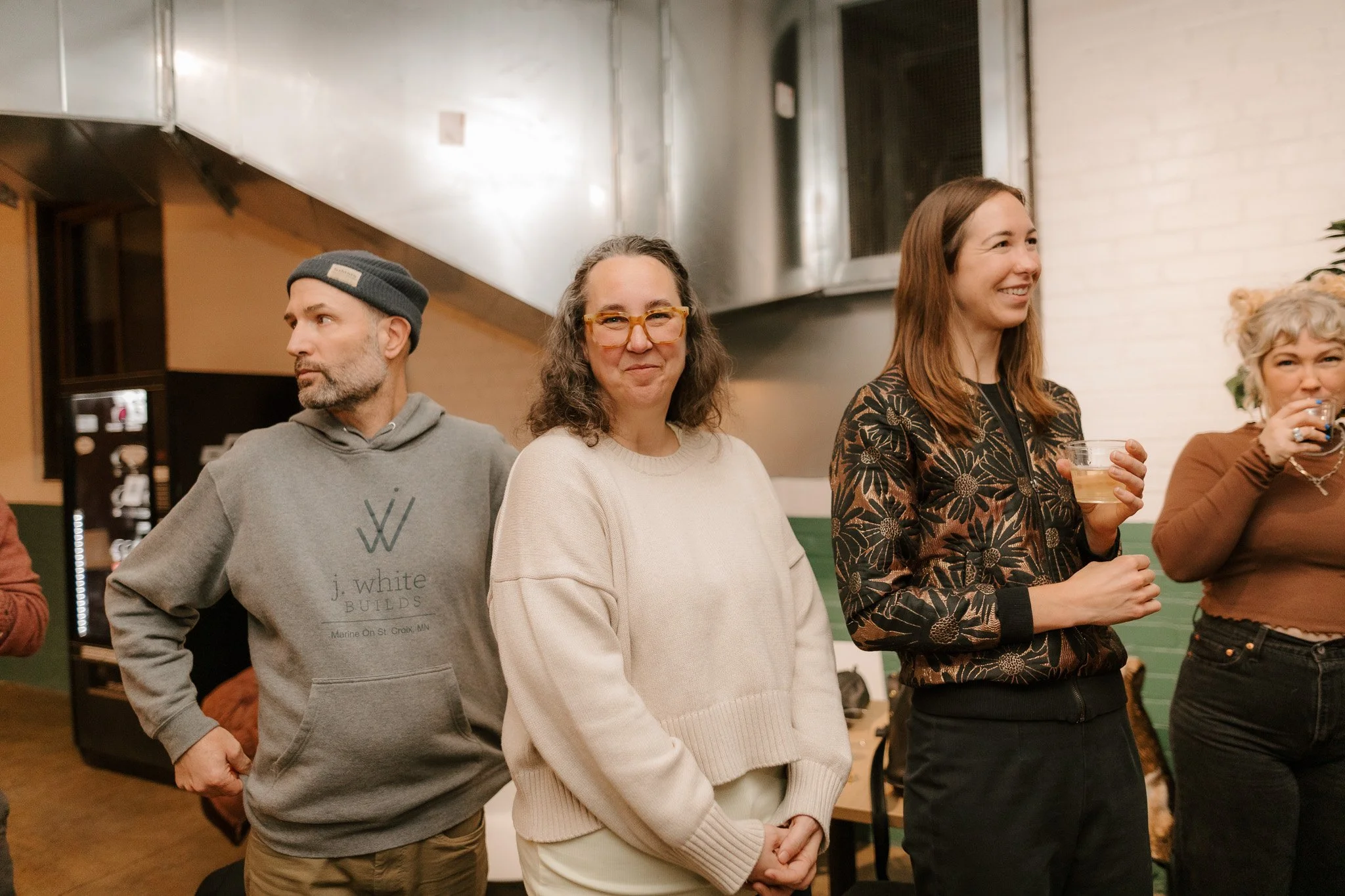 Four people standing in a row at a social gathering, indoors, with drinks in hand, smiling and engaging.