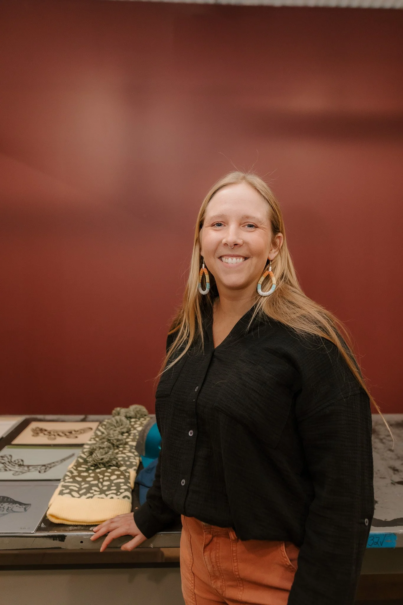 A woman with long blonde hair, smiling, wearing a black shirt and orange pants, standing in front of a display table with artwork and fabric samples, against a red wall.