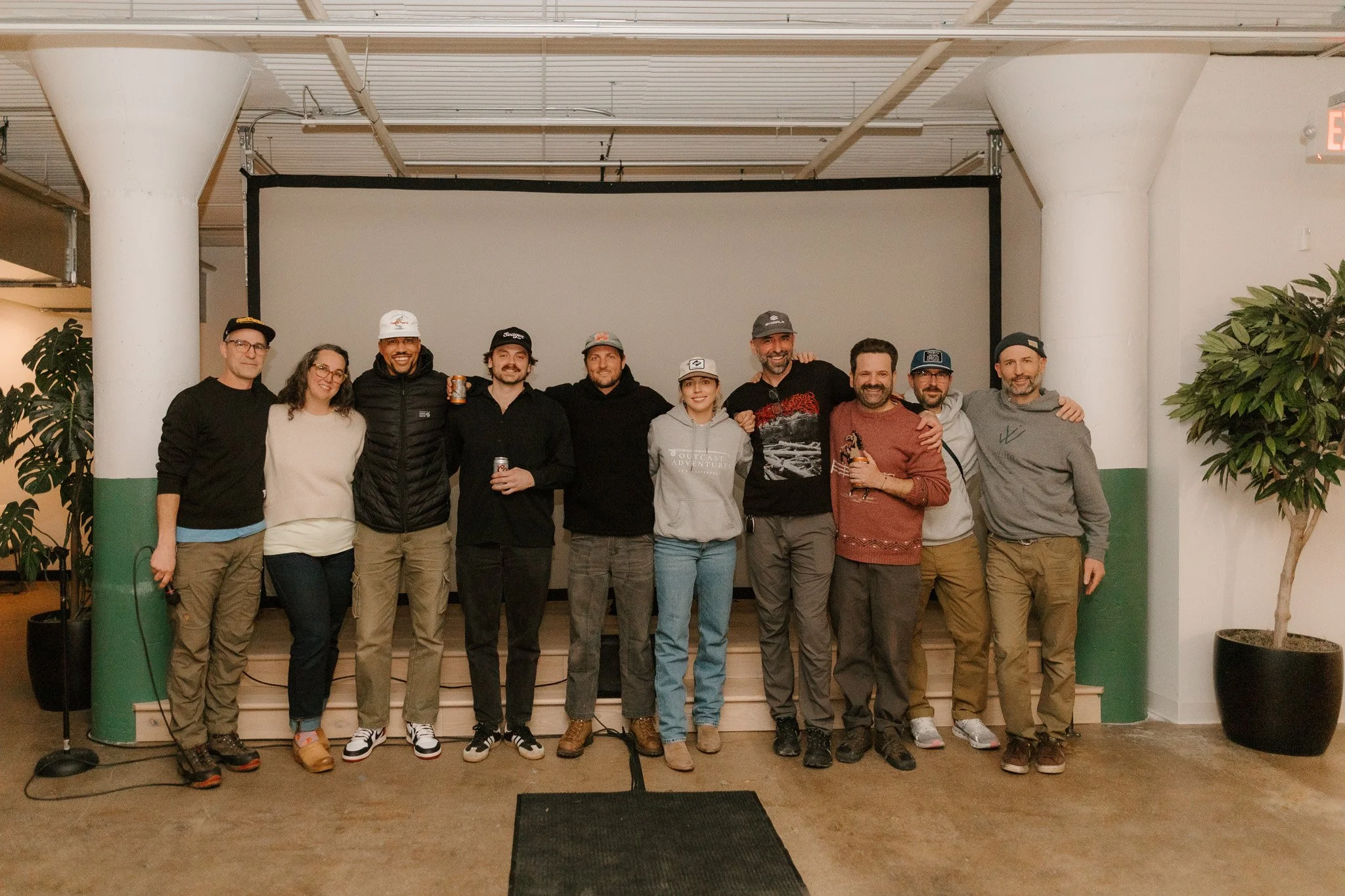 Group of eleven people posing together indoors in front of a large screen, with some holding drinks, smiling and standing arm in arm, possibly at a social or professional event.