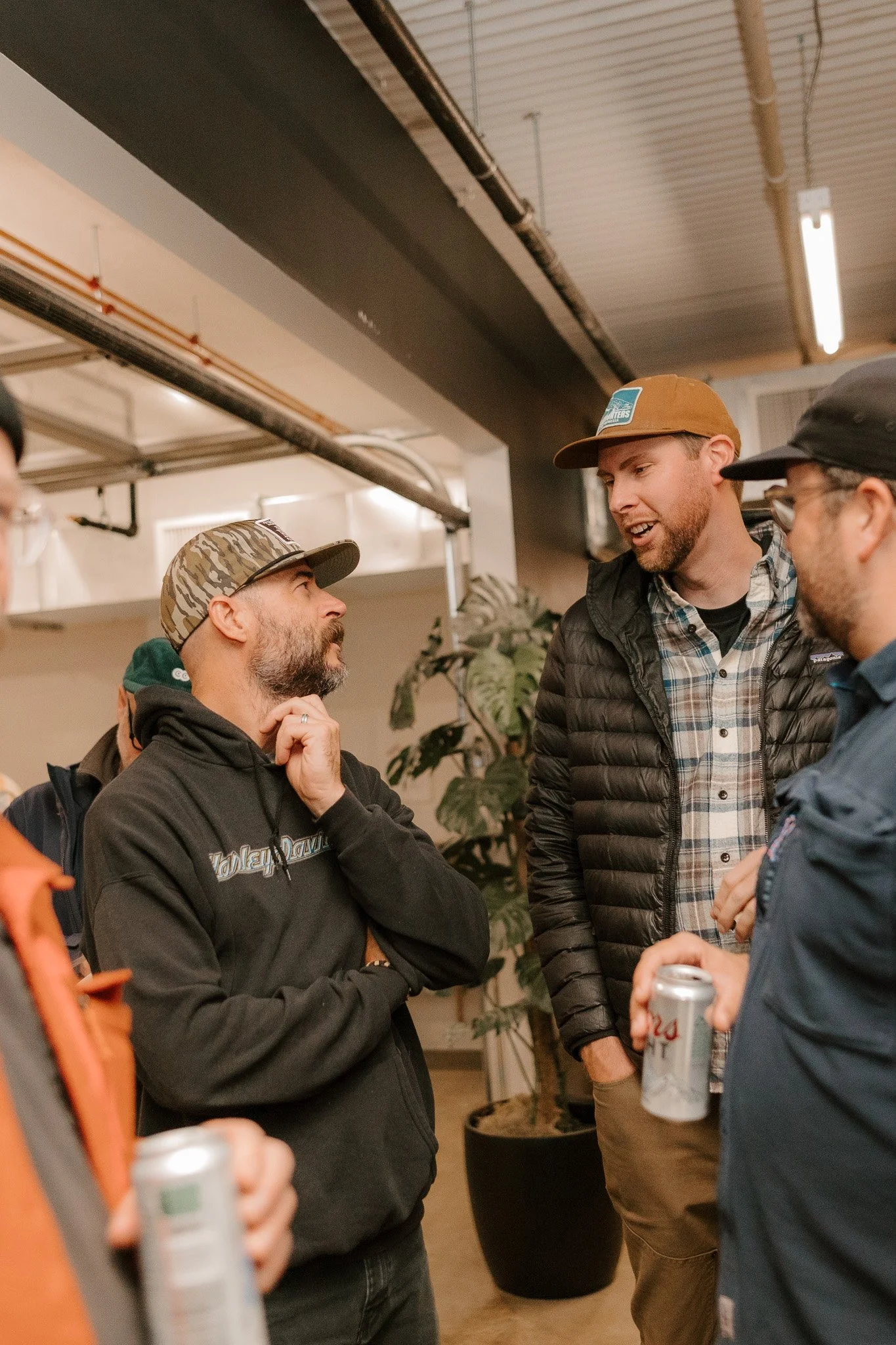Four men are having a conversation indoors, with one man holding a can of beer, in a casual setting with plants and industrial ceiling elements visible.