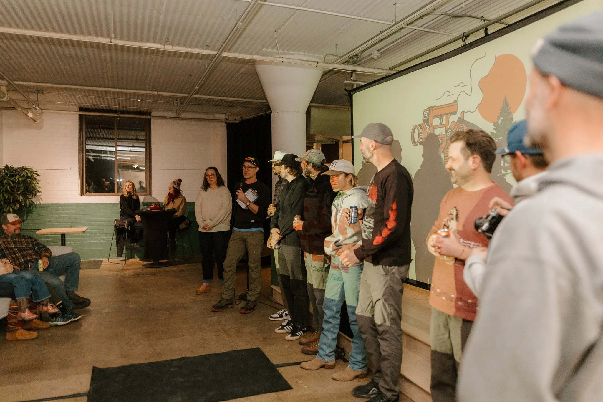 Group of people standing in a row on a stage, some holding drinks, at an indoor event in a spacious room with a projector screen displaying a cartoon of a camera, trees, and a sunrise or sunset in the background. Audience members are seated, watching