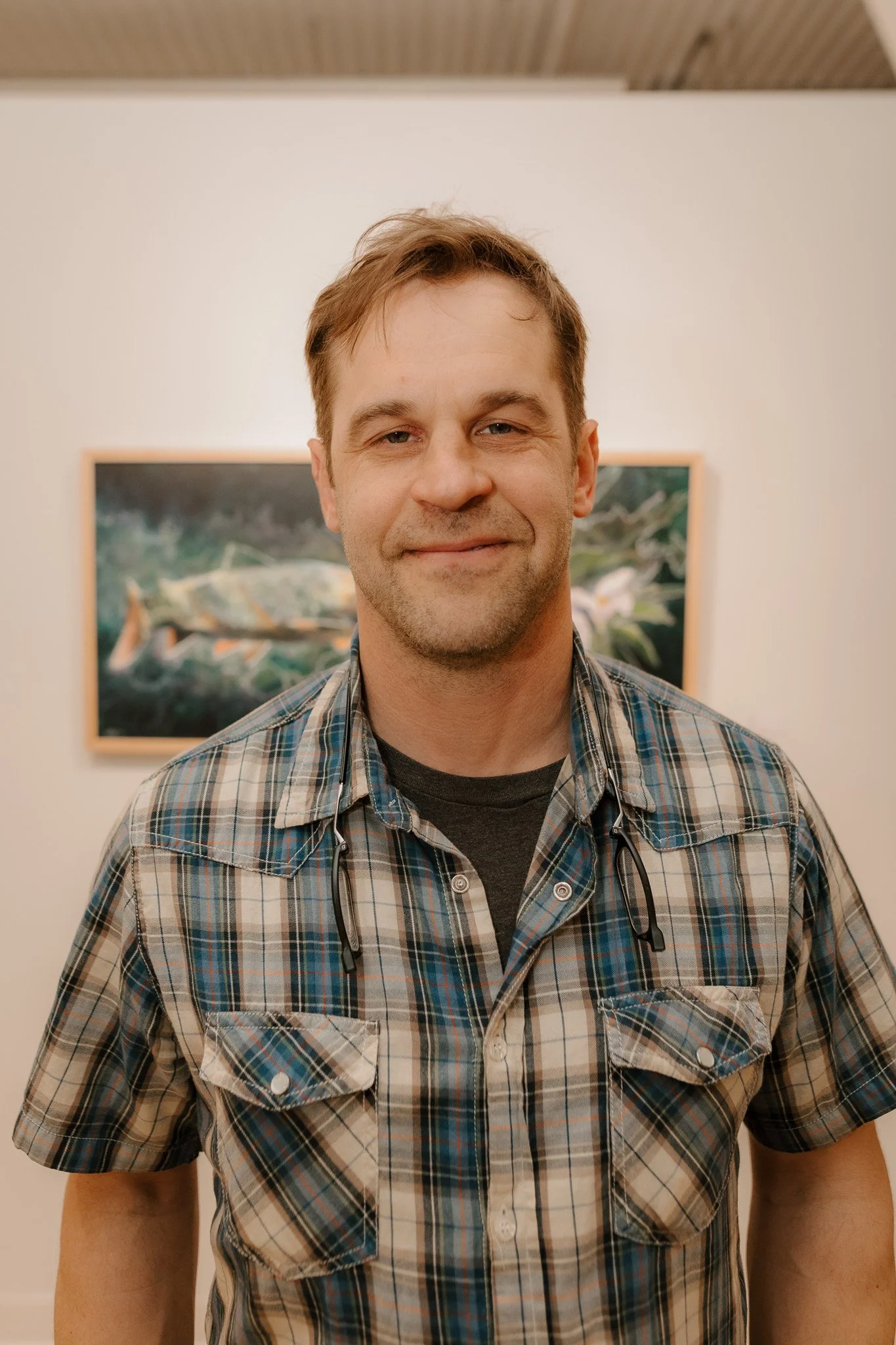 A man with short brown hair and a slight beard, smiling, standing indoors with framed artwork of fish in the background, wearing a plaid shirt and glasses hanging around his neck.