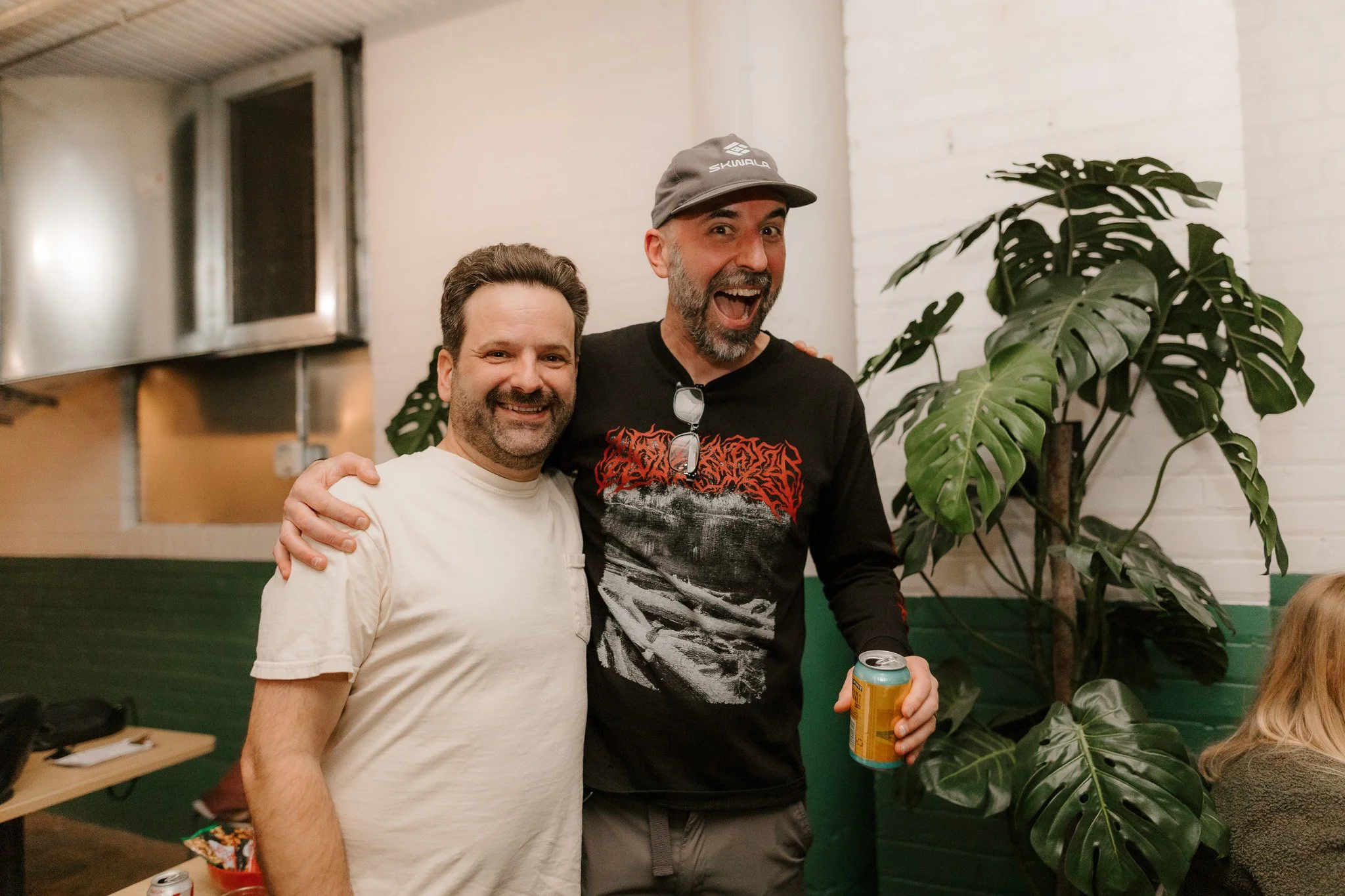 Two men smiling and posing together indoors, one wearing a light-colored t-shirt and the other in a black graphic long-sleeve shirt with a gray cap, holding a can of drink, with a large green plant behind them.
