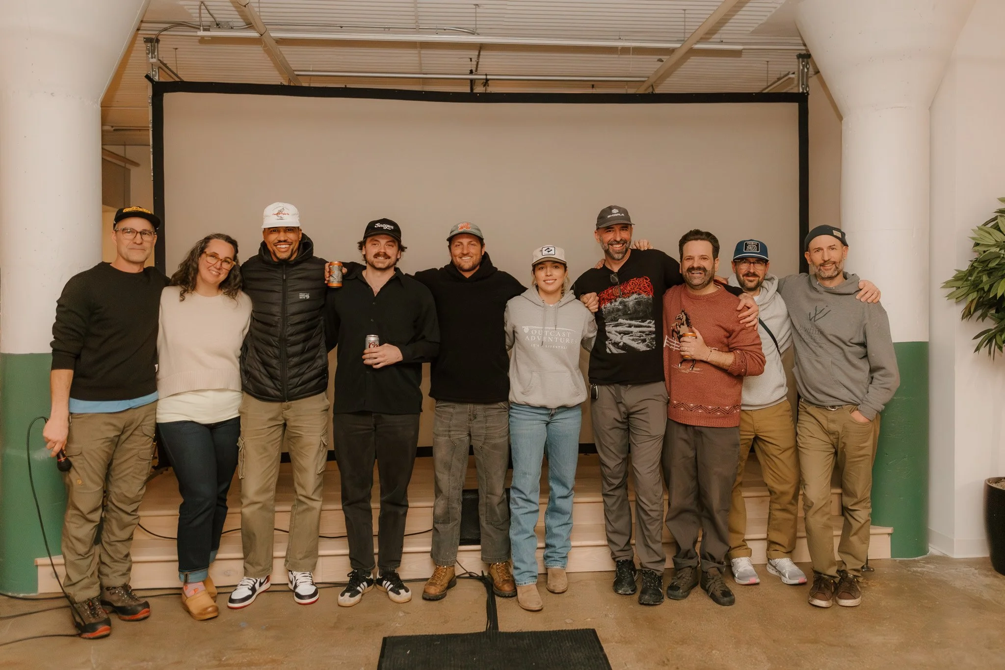 Group of twelve people standing together indoors, smiling, with a blank projection screen and white-painted columns in the background.