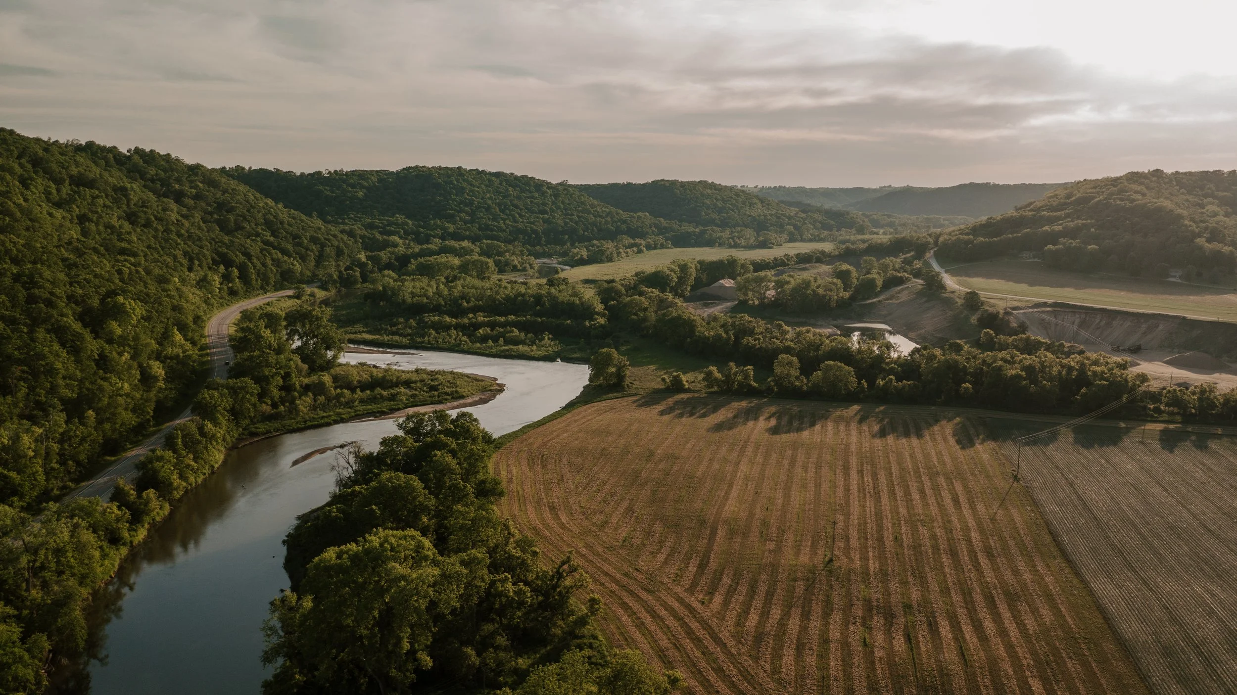 Aerial view of a river flowing through green forested hills and farmland, with overcast skies.