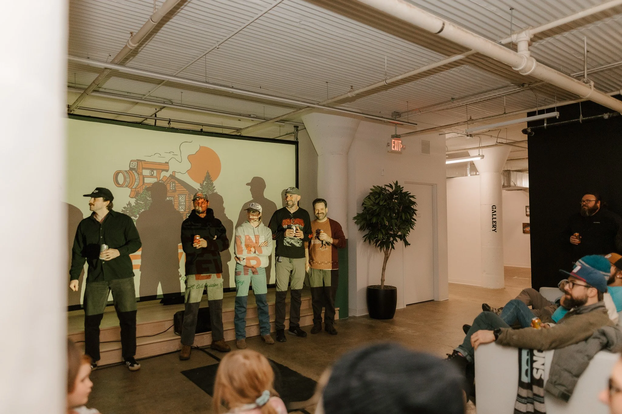 Group of people standing on a stage during an event, some holding drinks, with a projection of a house and trees in the background.