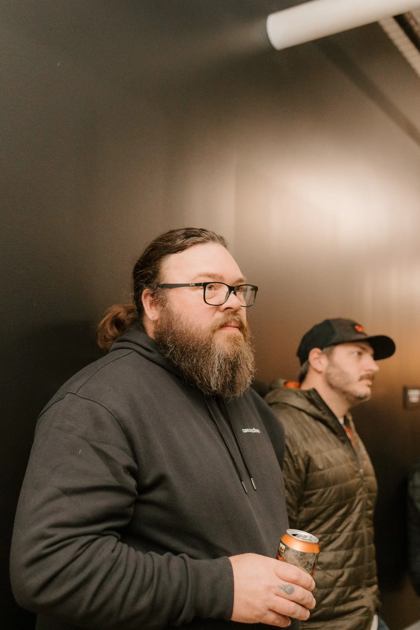 A bearded man with glasses wearing a black hoodie holding a can of beer at a gathering, with another man in a cap and brown jacket in the background, against a dark wall.