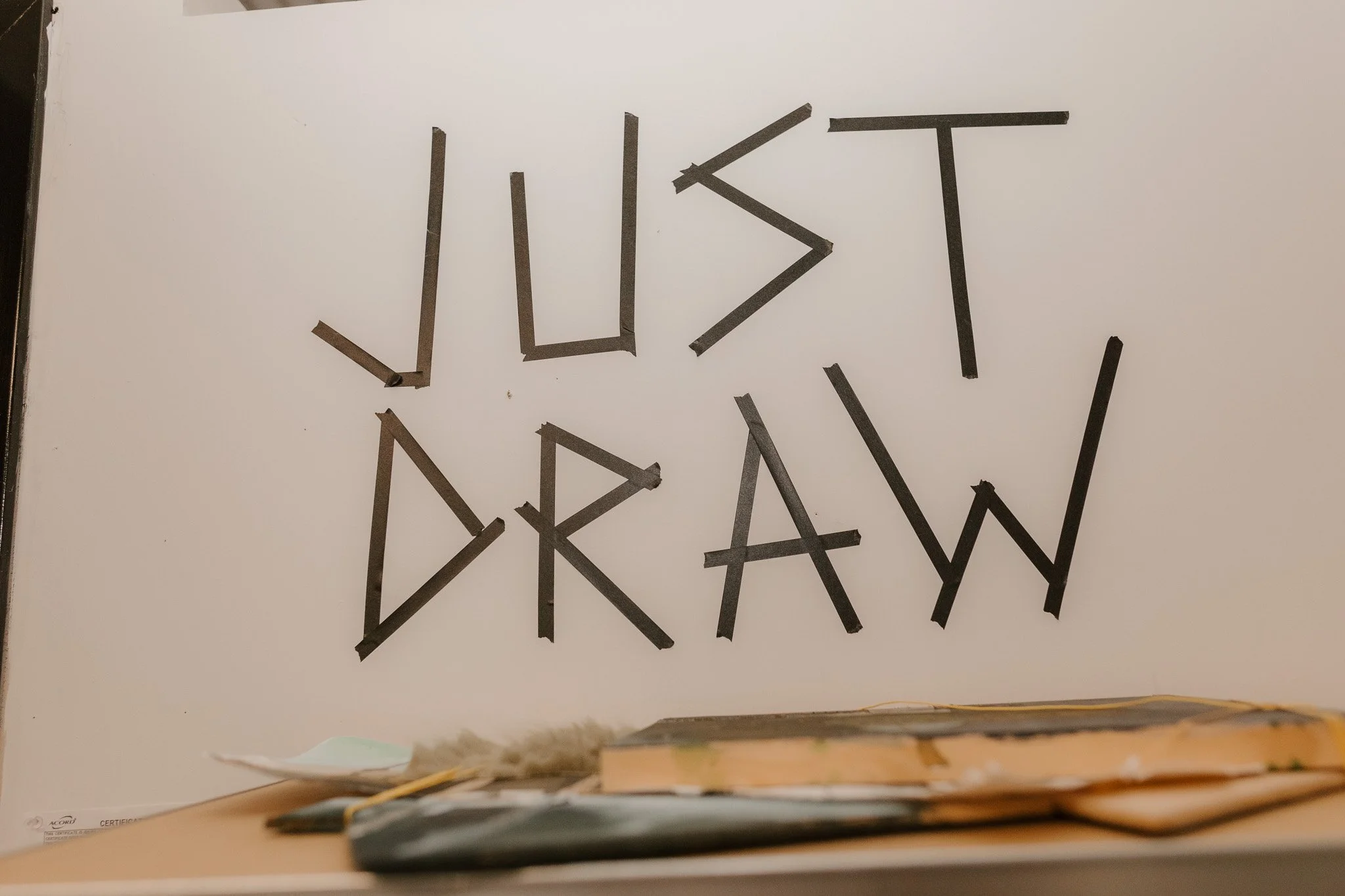 Letters spelling 'JUST DRAW' formed with black tape on a whiteboard