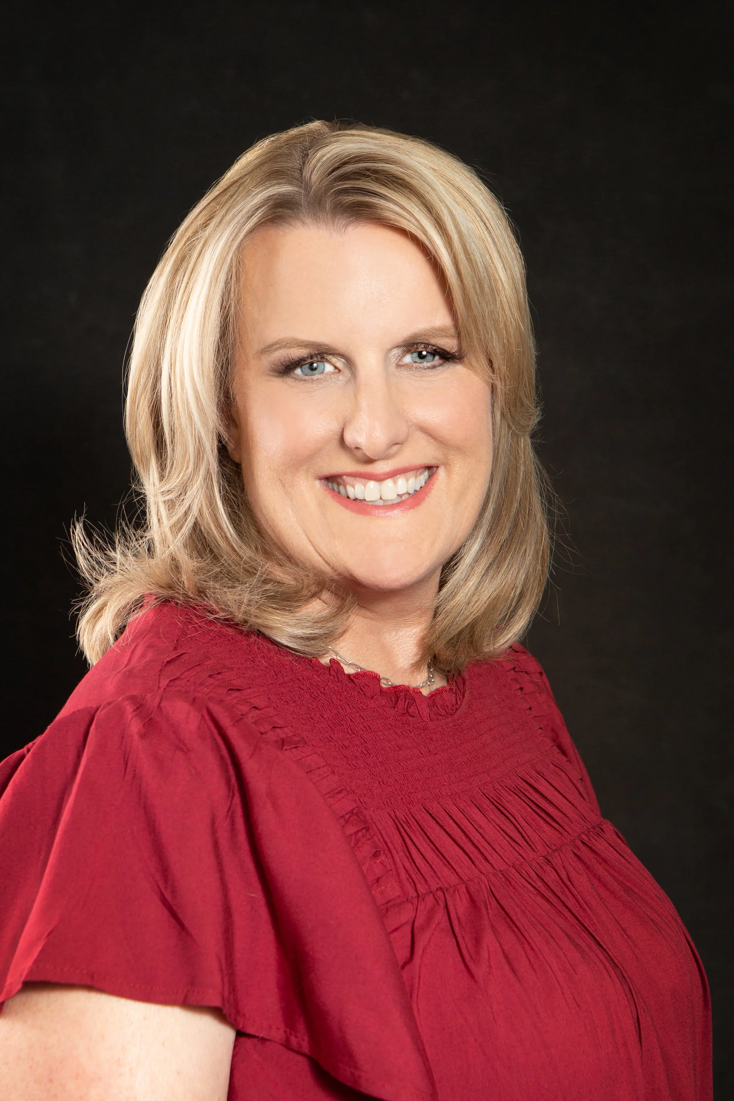 Portrait of a smiling woman with blonde hair wearing a red blouse against a black background.