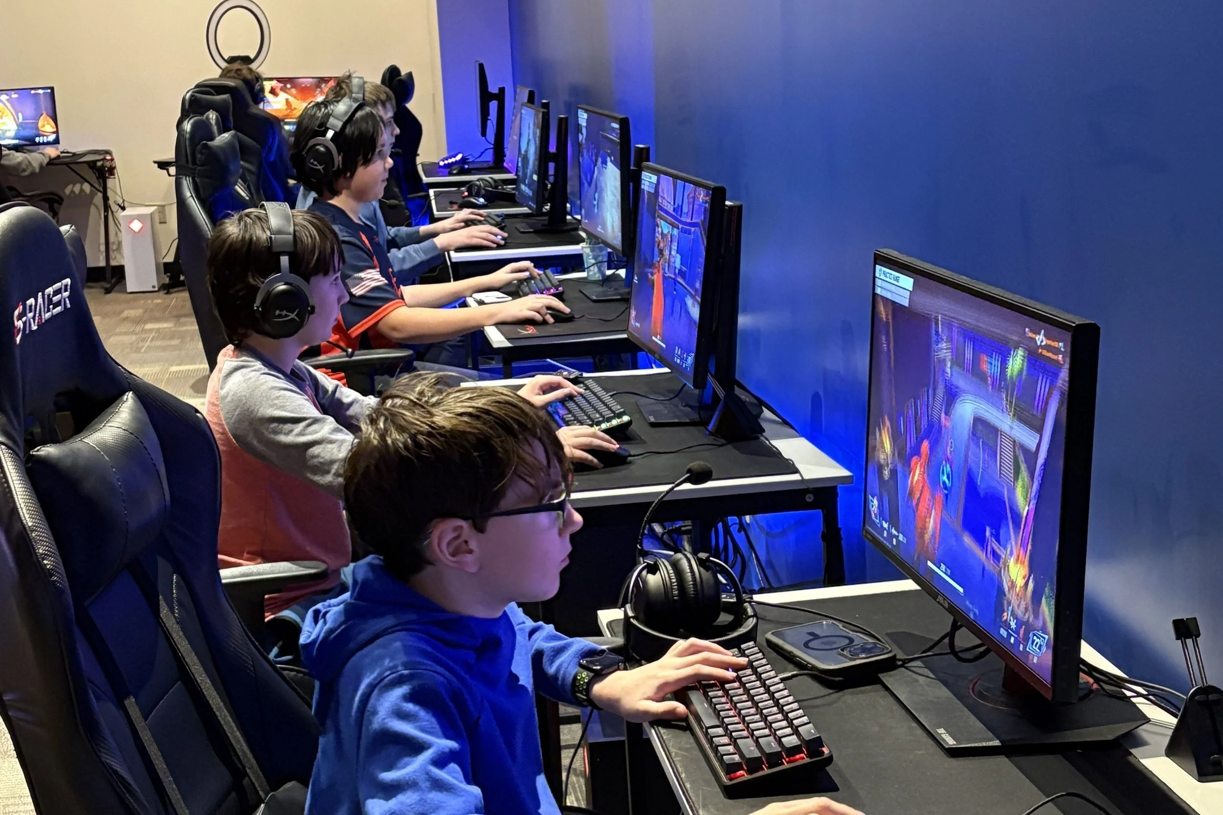 Several children are sitting in a row at computer stations, playing video games in a gaming center. They are wearing headphones and focused on their screens, with colorful game visuals displayed.
