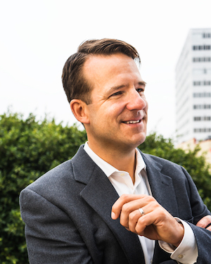 Smiling man in a gray suit outdoors with green bushes and a modern office building in the background.