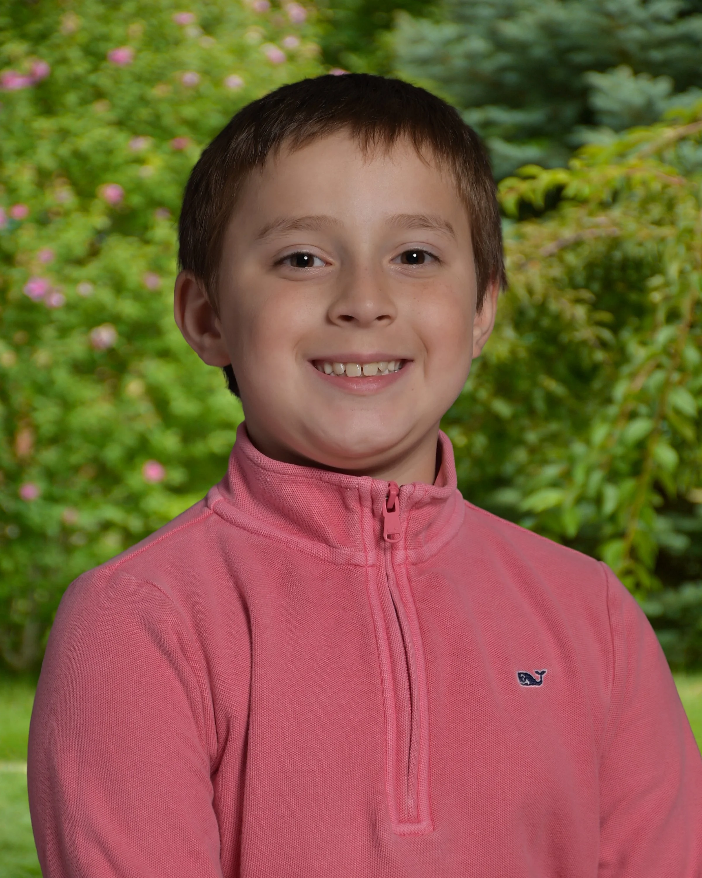 A young boy with short brown hair, smiling and wearing a pink zip-up jacket, standing outdoors with a background of green trees and pink flowers.