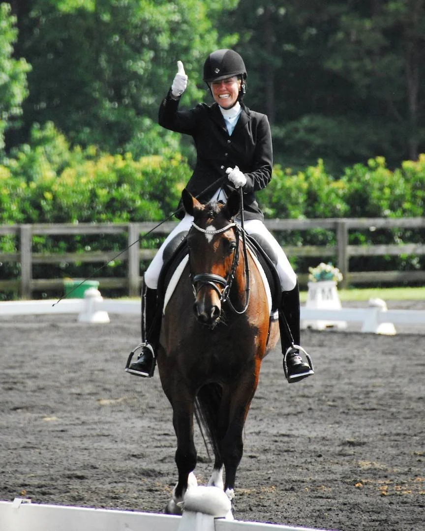 A woman in riding attire riding a brown horse during a equestrian event, smiling and waving, outdoors on a dirt arena with green trees in the background.