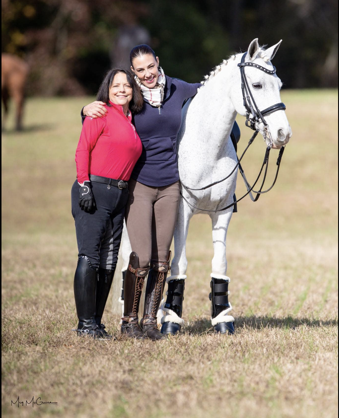 Two women standing outdoors with a white horse, smiling, one with arm around the other's shoulder, both dressed in equestrian attire.