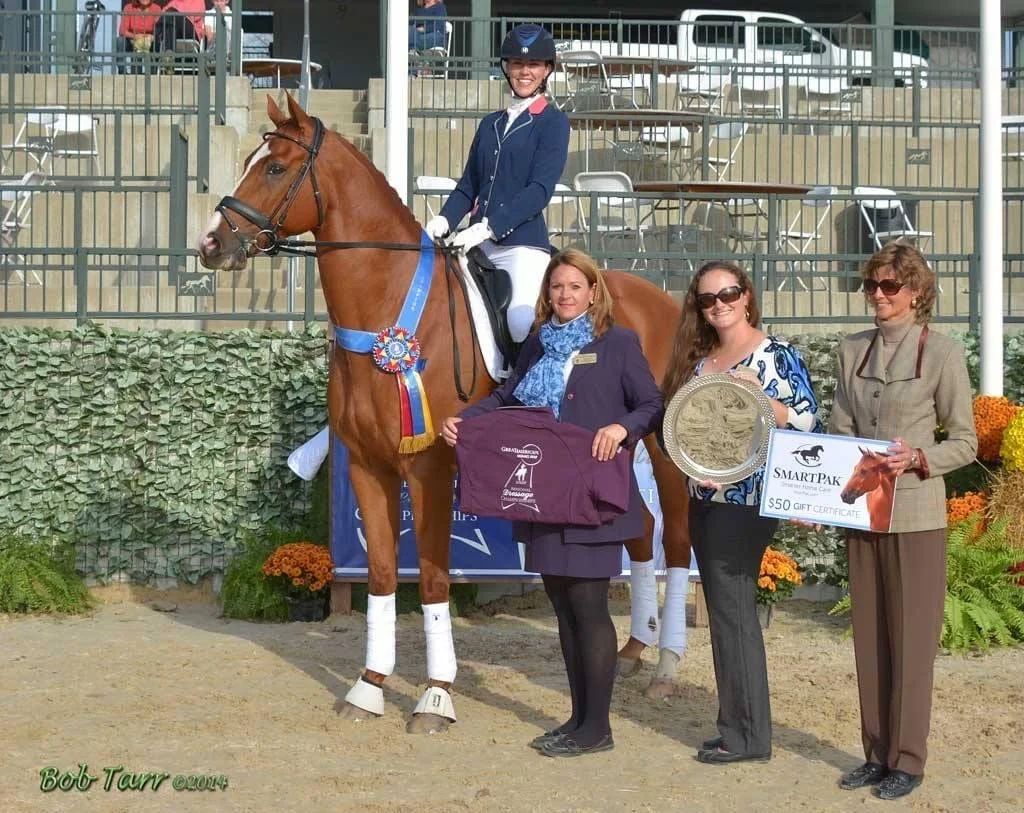 A woman in riding attire on a horse with a blue ribbon, surrounded by three women holding awards and a gift certificate, in an outdoor arena setting.
