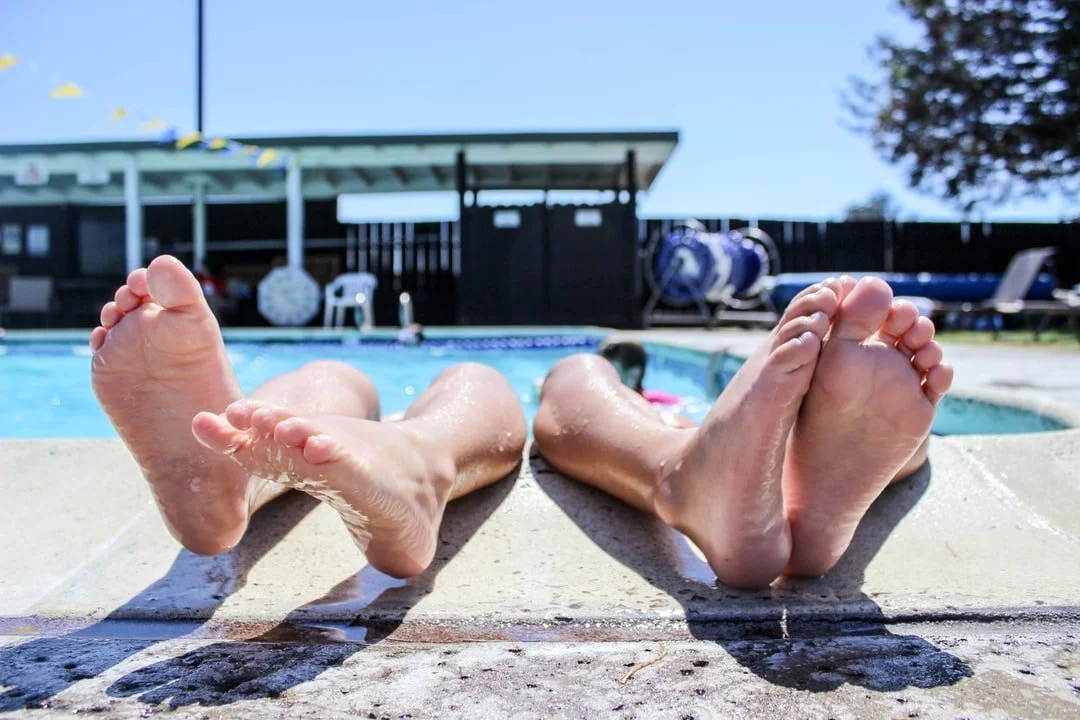 Two people relaxing by a swimming pool with their feet stretched out on the poolside, sunny day, backyard setting.