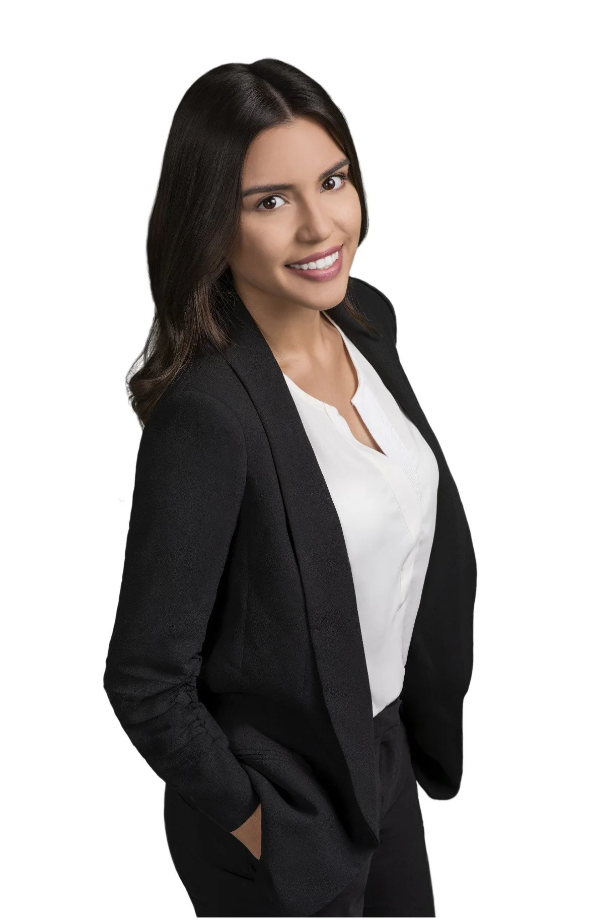 A woman in a black blazer and white blouse with long dark hair smiling, posing with her hands in her pockets on a white background.