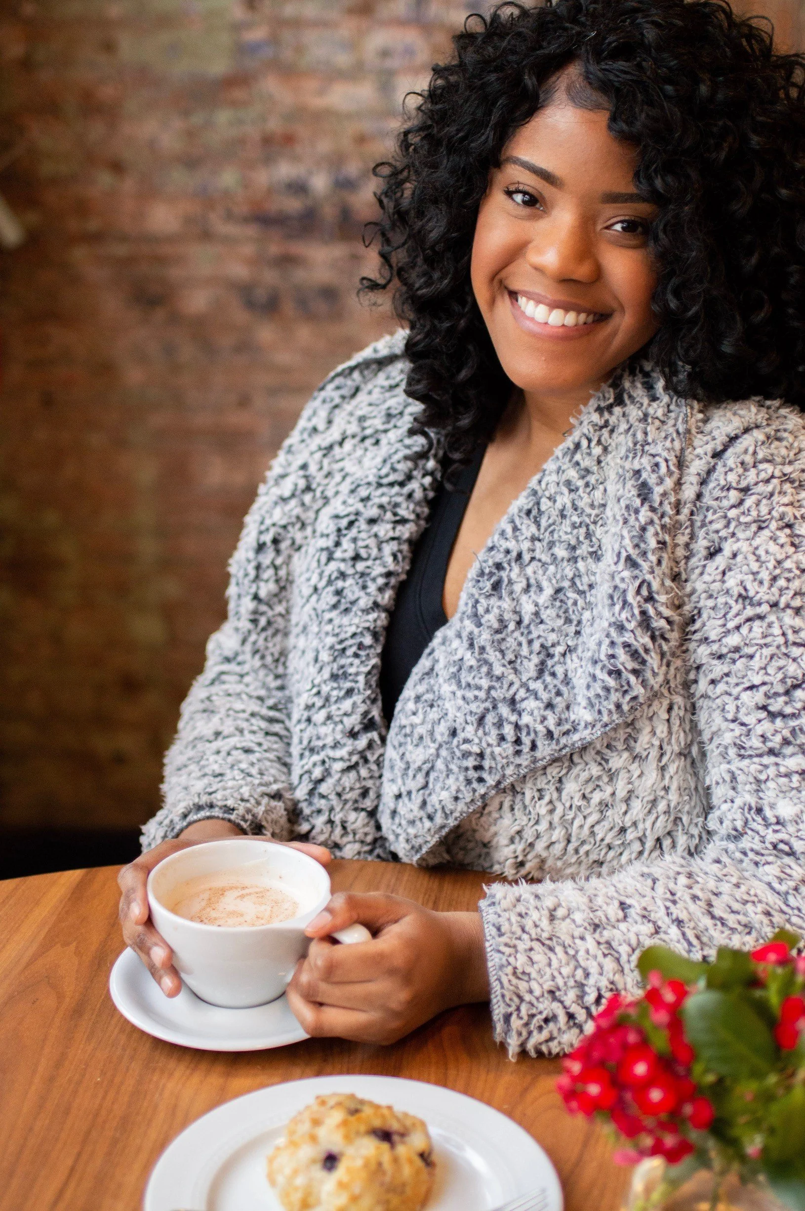 A woman with curly black hair sitting at a table, smiling and holding a cup of coffee or hot chocolate in a white cup on a saucer. On the table in front of her, there is a plate with a cookie, and in the foreground, there is a blurry vase with pink flowers. The background shows a brick wall.