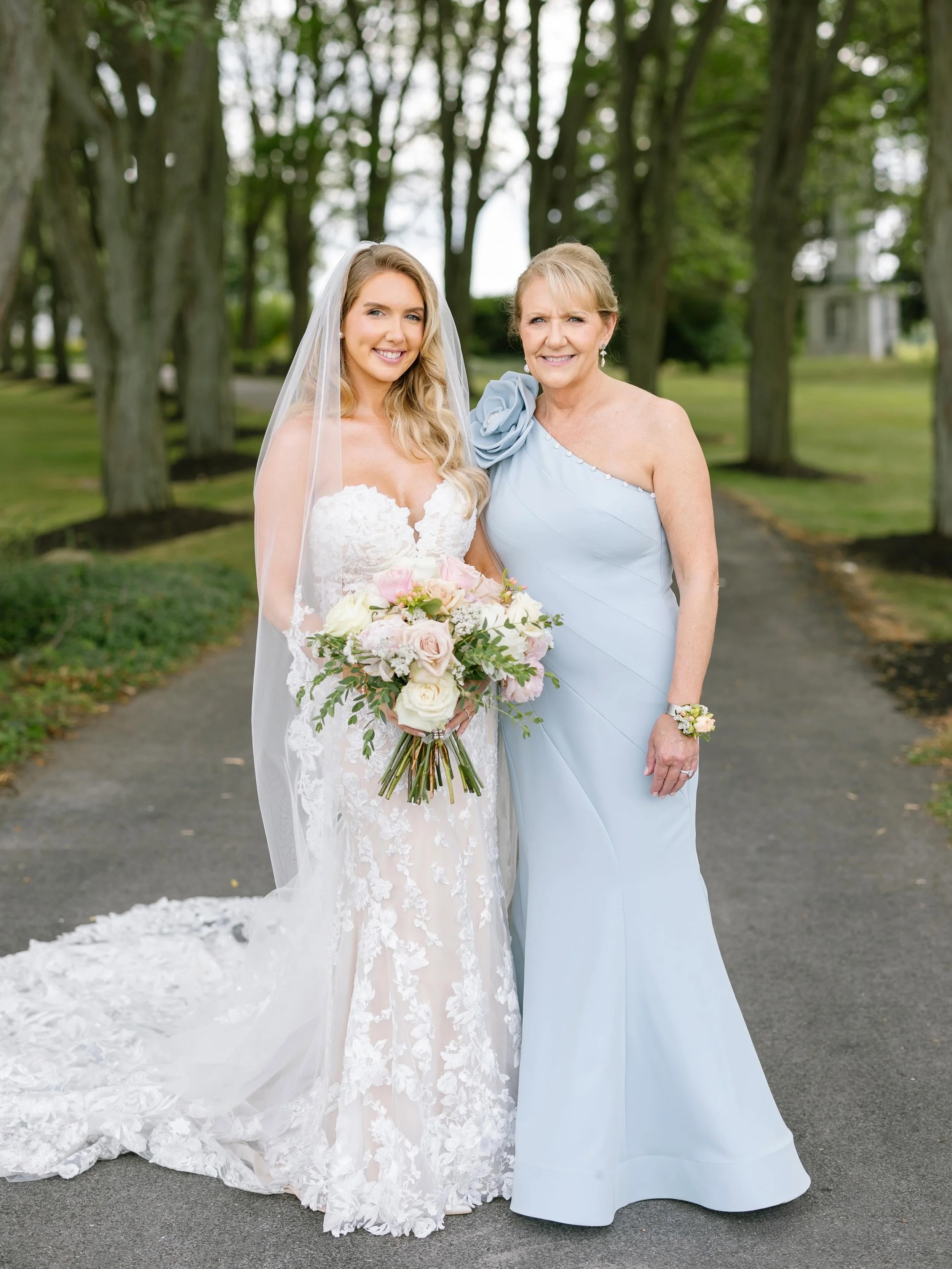 Mother of the bride and bride standing side by side in a park. Mother of the bride wearing one shoulder gown with 3d flower at shoulders in powder blue by Mac Duggal