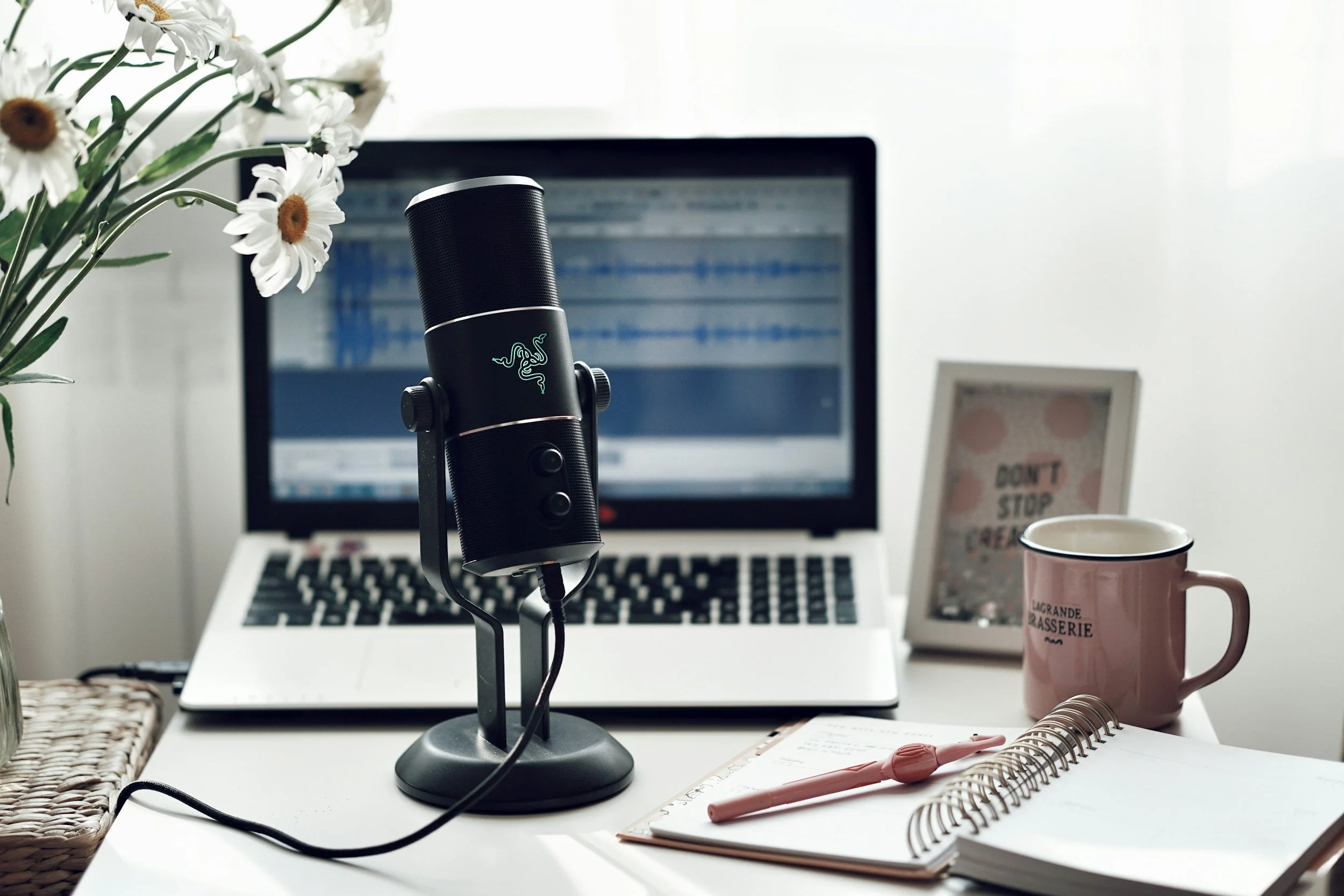 laptop with notebook coffe mug and picture frame to the right, microphone in front and daisies in left corner