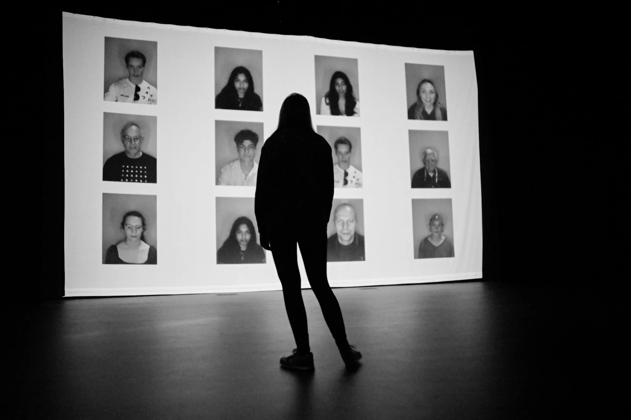 Silhouette of a person standing in front of a large screen displaying multiple black and white portrait photos.
