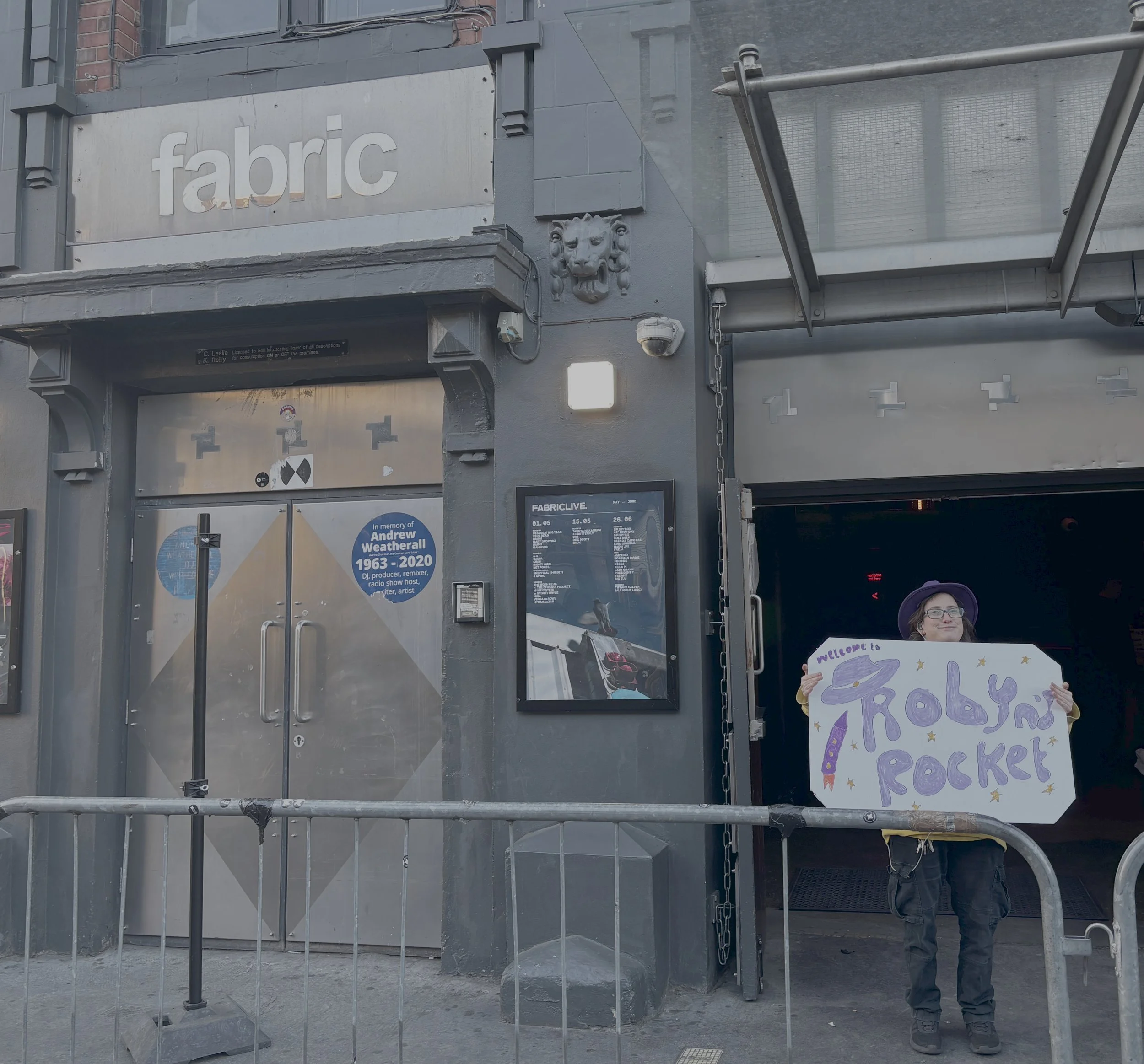 a building with the word fabric above it, a person stands outside holding a sign which reads Robyn's Rocket with a hat on the first R, a cartoon rocket and stars.