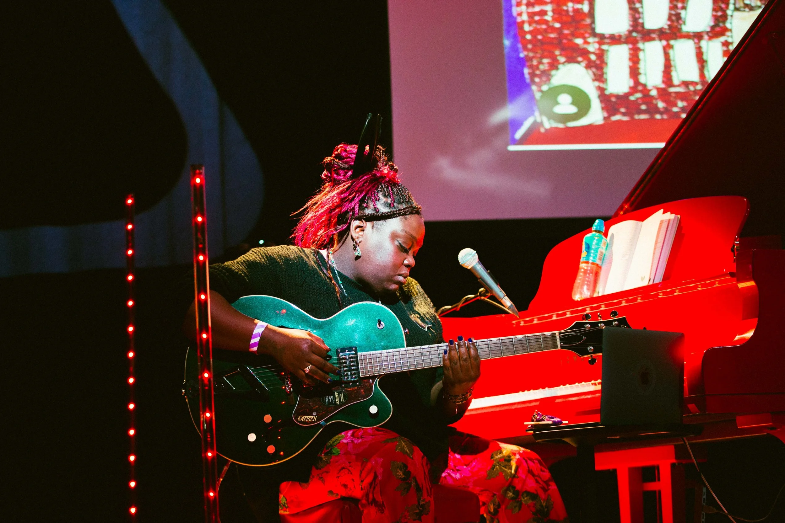 Donna Thompson playing a blue guitar , sat at a red piano , with red lights behind her , and part of a screen visible with a hand drawn  picture of the royal Albert hall  Photo by Sian O Connor.