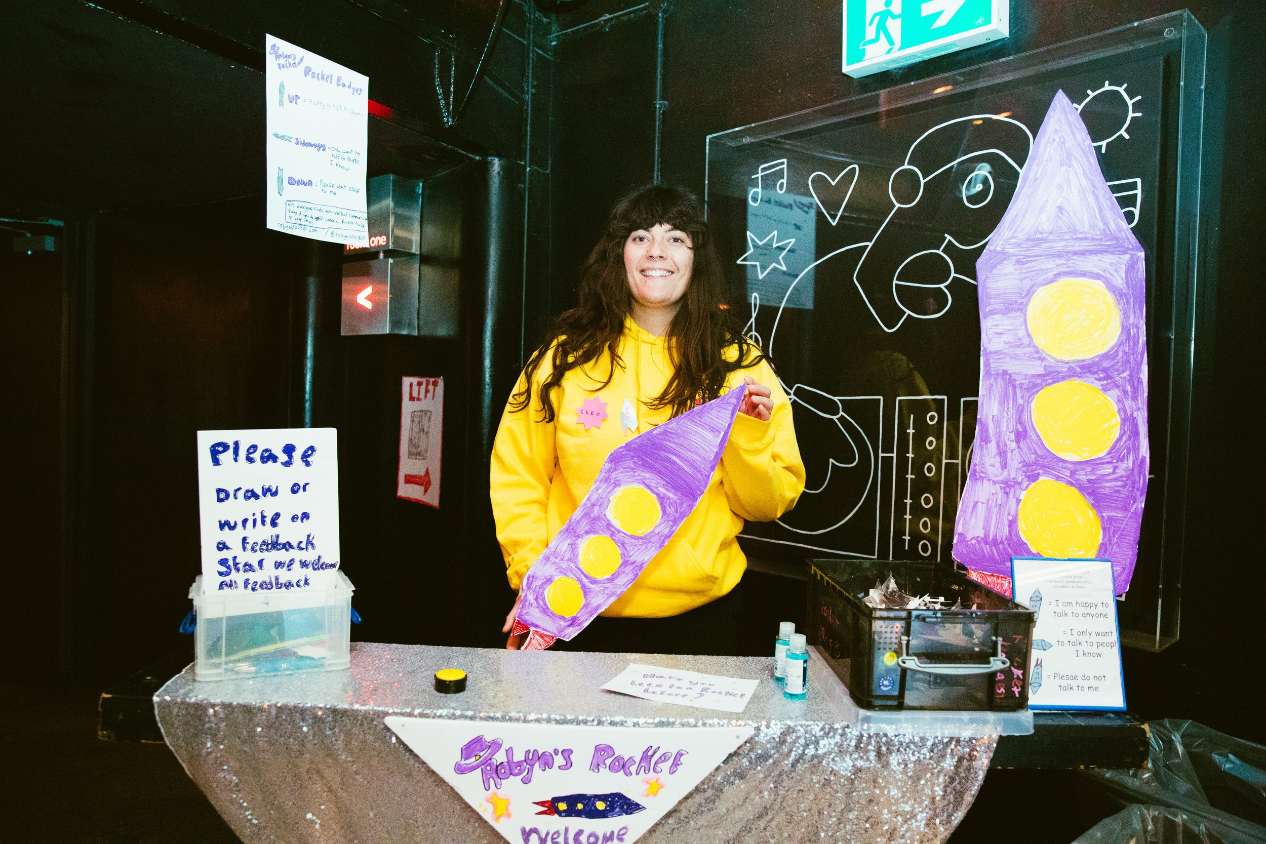 a white woman with shoulder length dark hair, holding a purple rocket made out of foamboard.