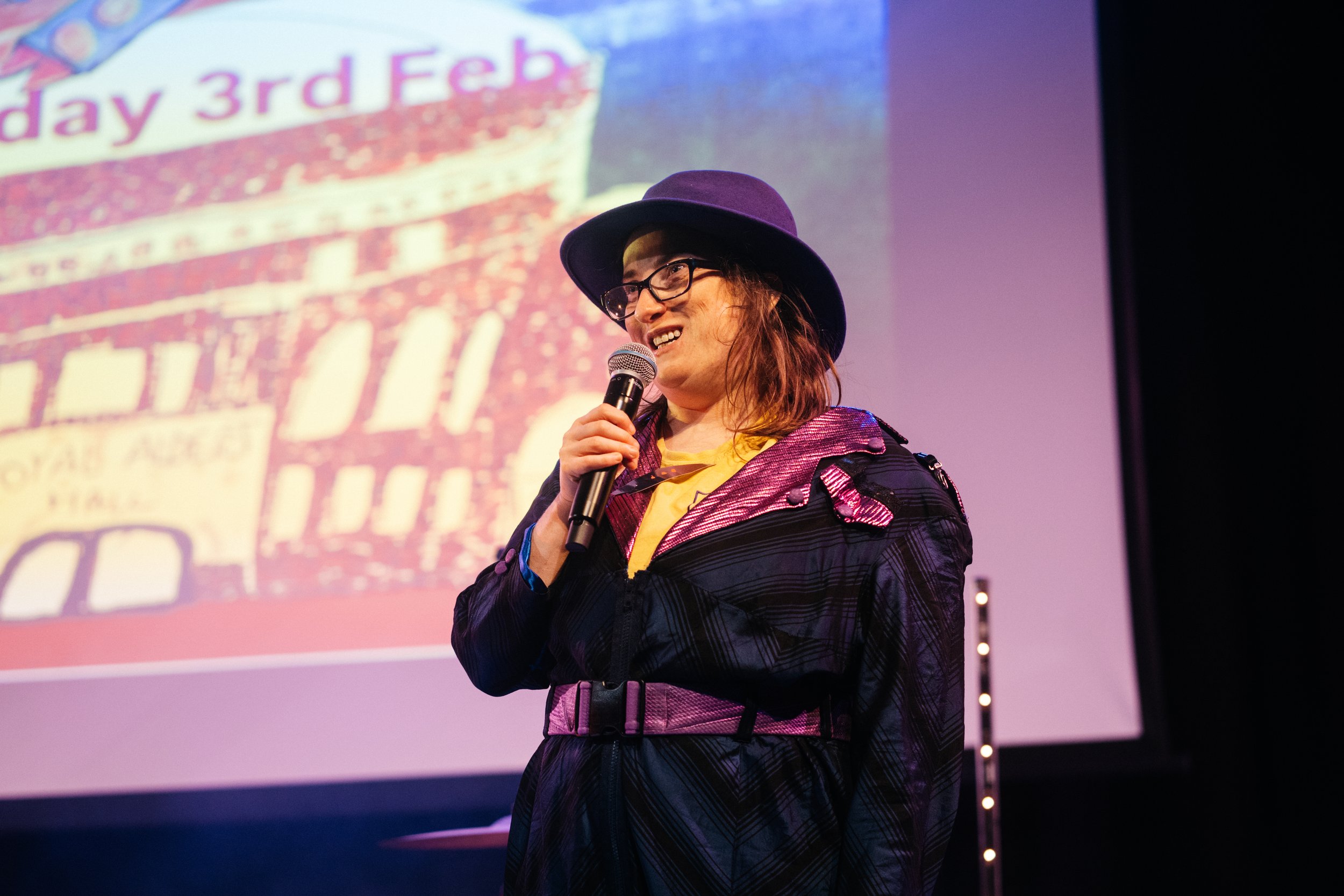 Robyn Rocket in a purple hat , blue and purple shiny boiler suit and yellow T shirt , speaking into a microphone on stage, with a projection of a hand drawn image of the royal Albert hall behind her Temi Adegbayibi 