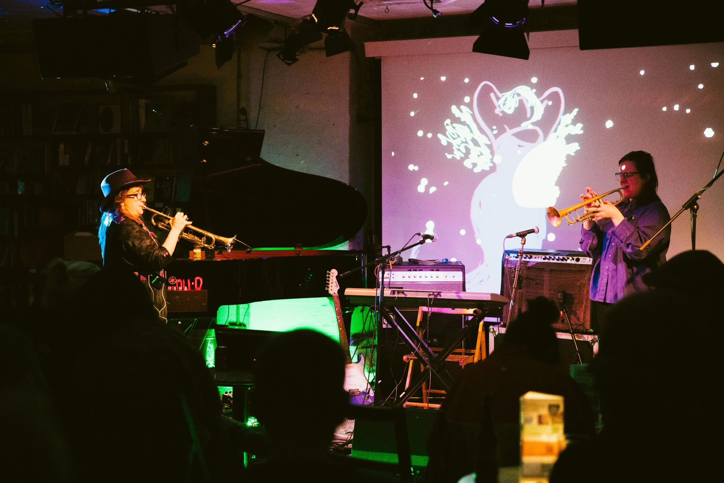 2 female bodied people on a flat stage with visuals behind them playing trumpets