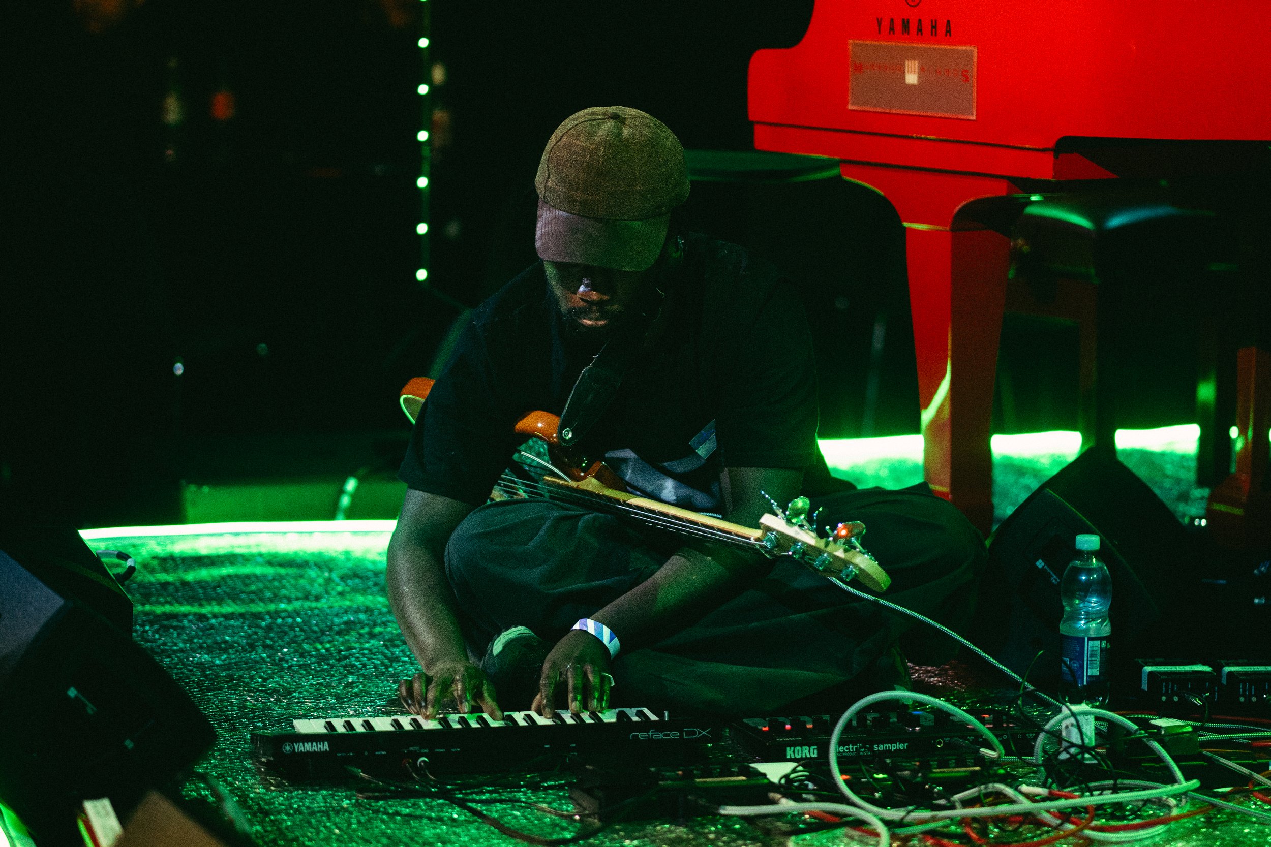 Coby Sey crouching playing keyboard and wearing a. bass guitar on a silver stage with green light  photo by Temi Adegbayibi 