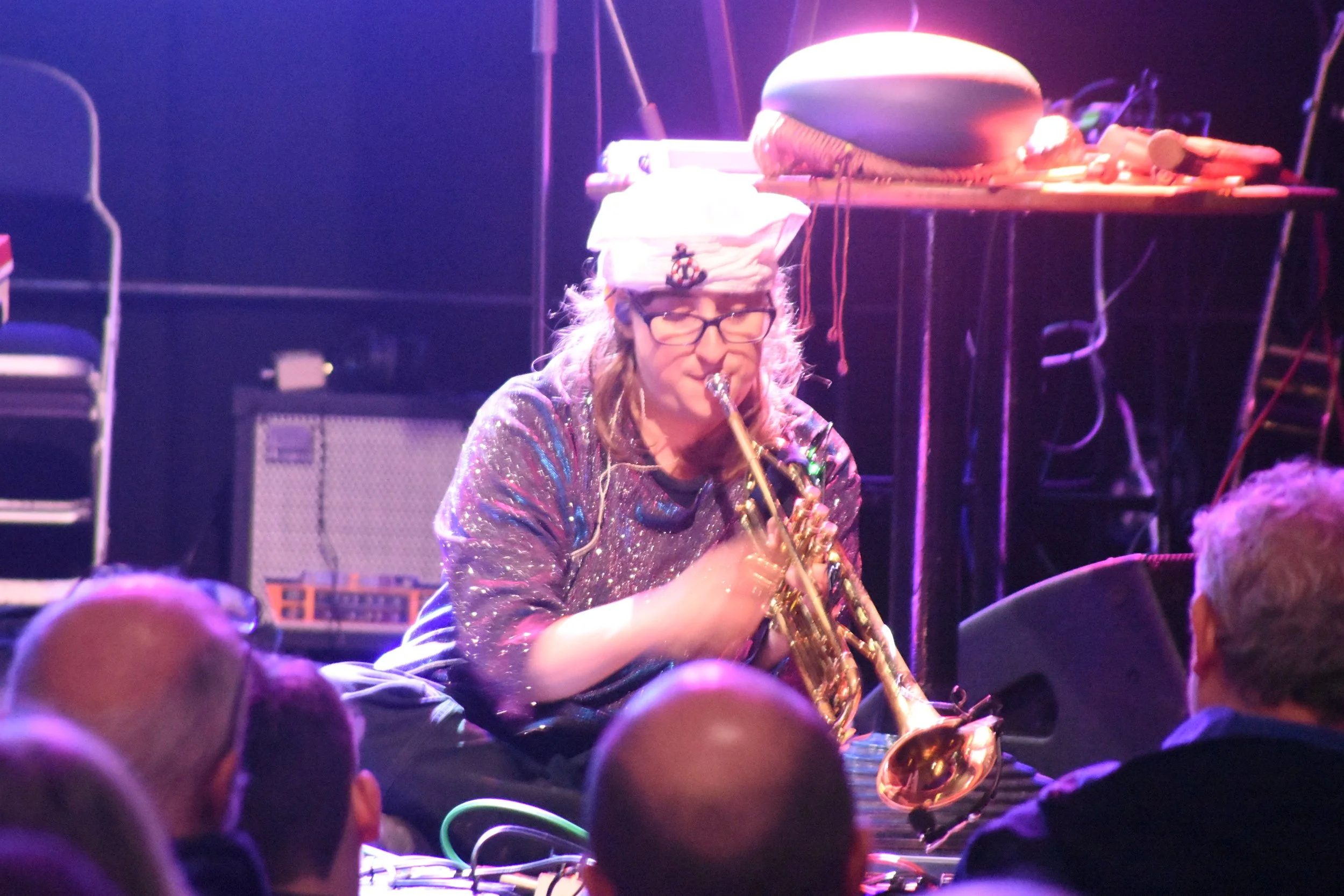 a white woman wearing a sailor hat and sparkly top sat on a stage with a trumpet and