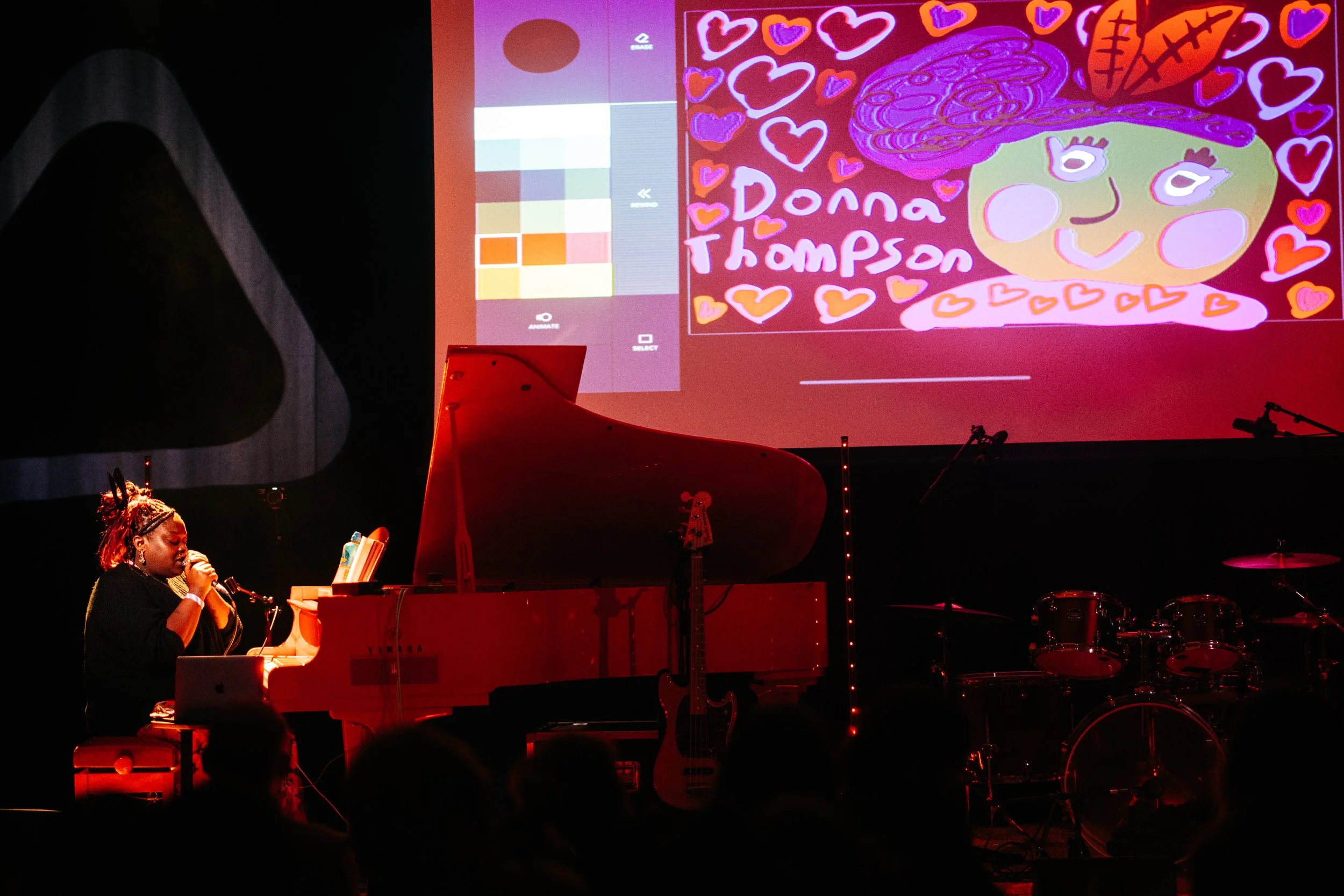Donna Thompson sat at a red piano in front of a screen with hand drawn picture of her. photo by Temi Adegbayibi 
