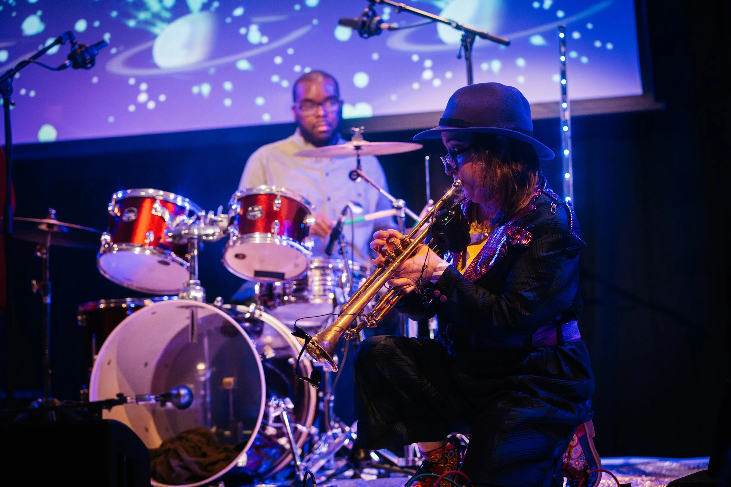 Robyn Rocket crouching playing Trumpet with Isaac Palm playing drums, with a projection screen with planets and stars on it. photo by Temi Adegbayibi 