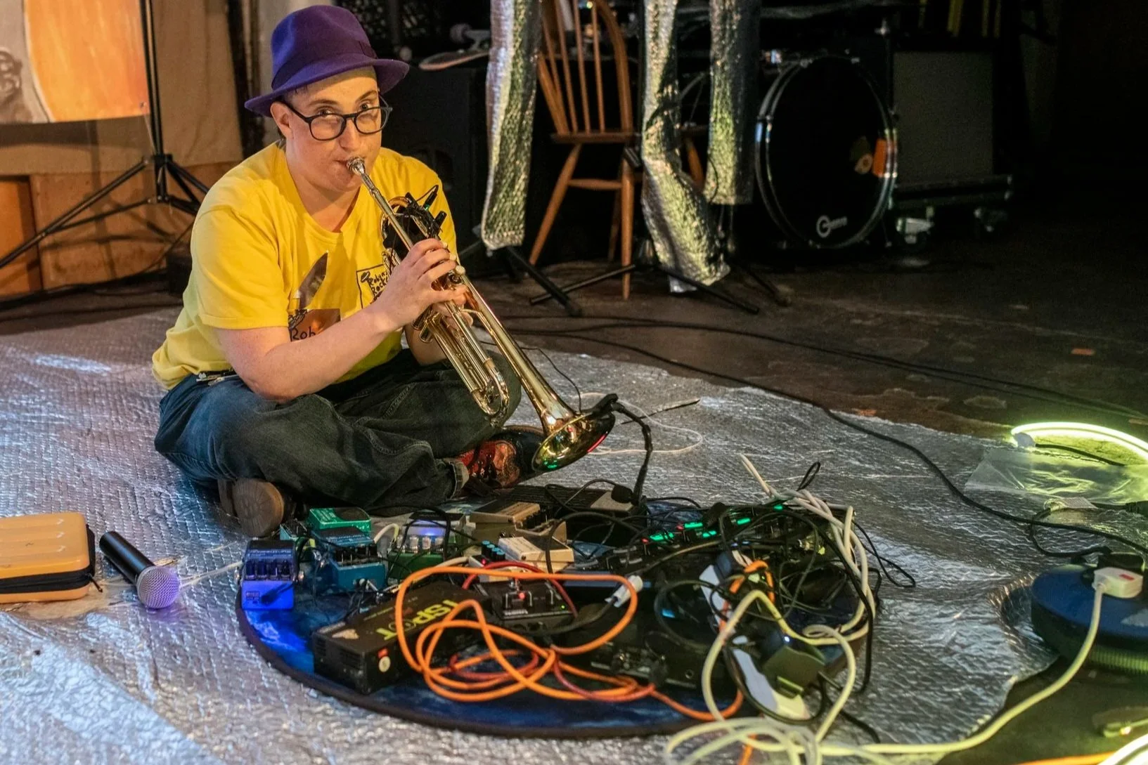 a white woman wearing a purple hat and glasses, sat on the floor , playing a trumpet through guitar pedals