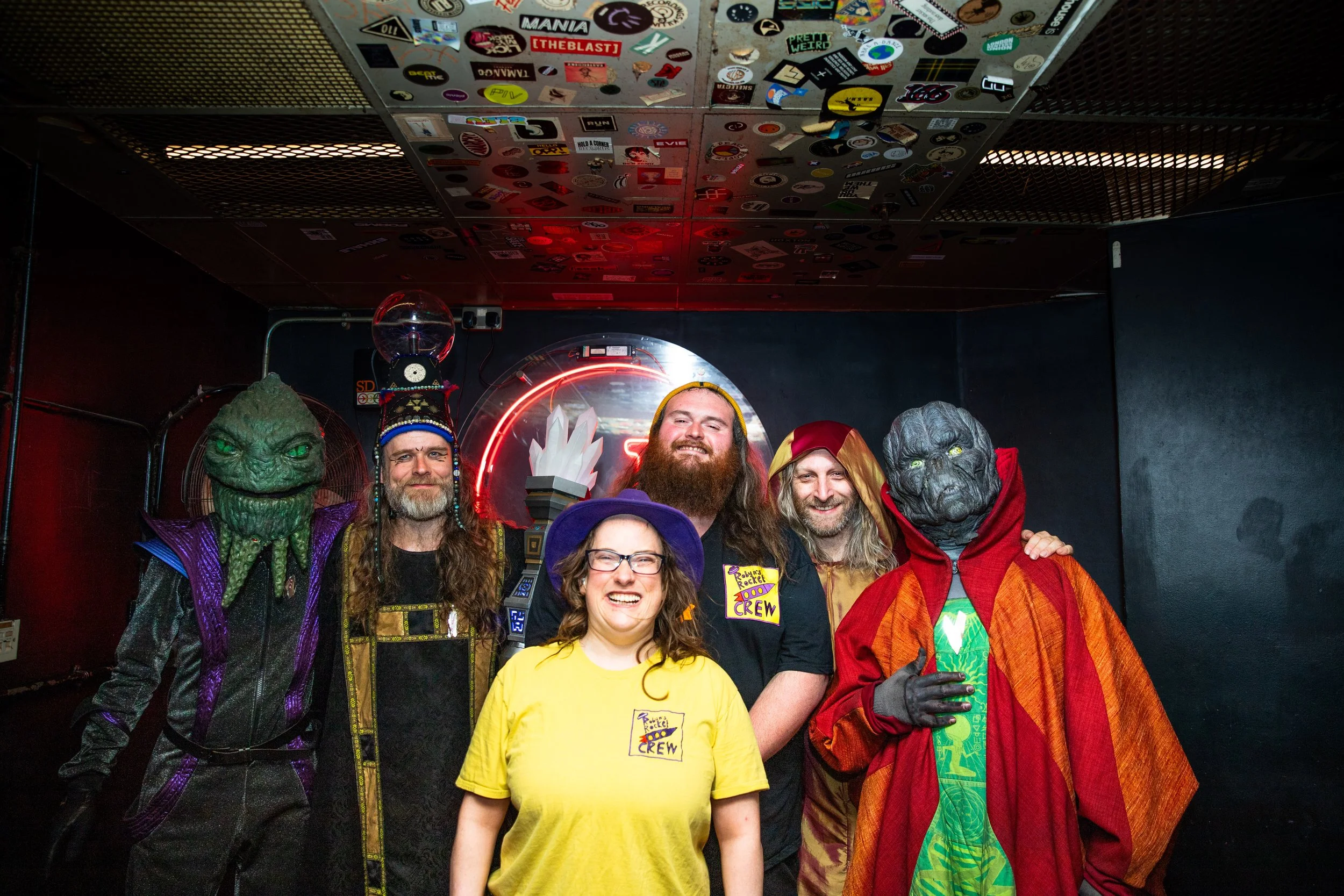 a photo of a green headed alien with tentacles under their face in a jump suit, a human looking alien with a sphere on their head, a white woman with a purple hat , a white man with shoulder length hair, a white man wearing a. robe, and a alien.
