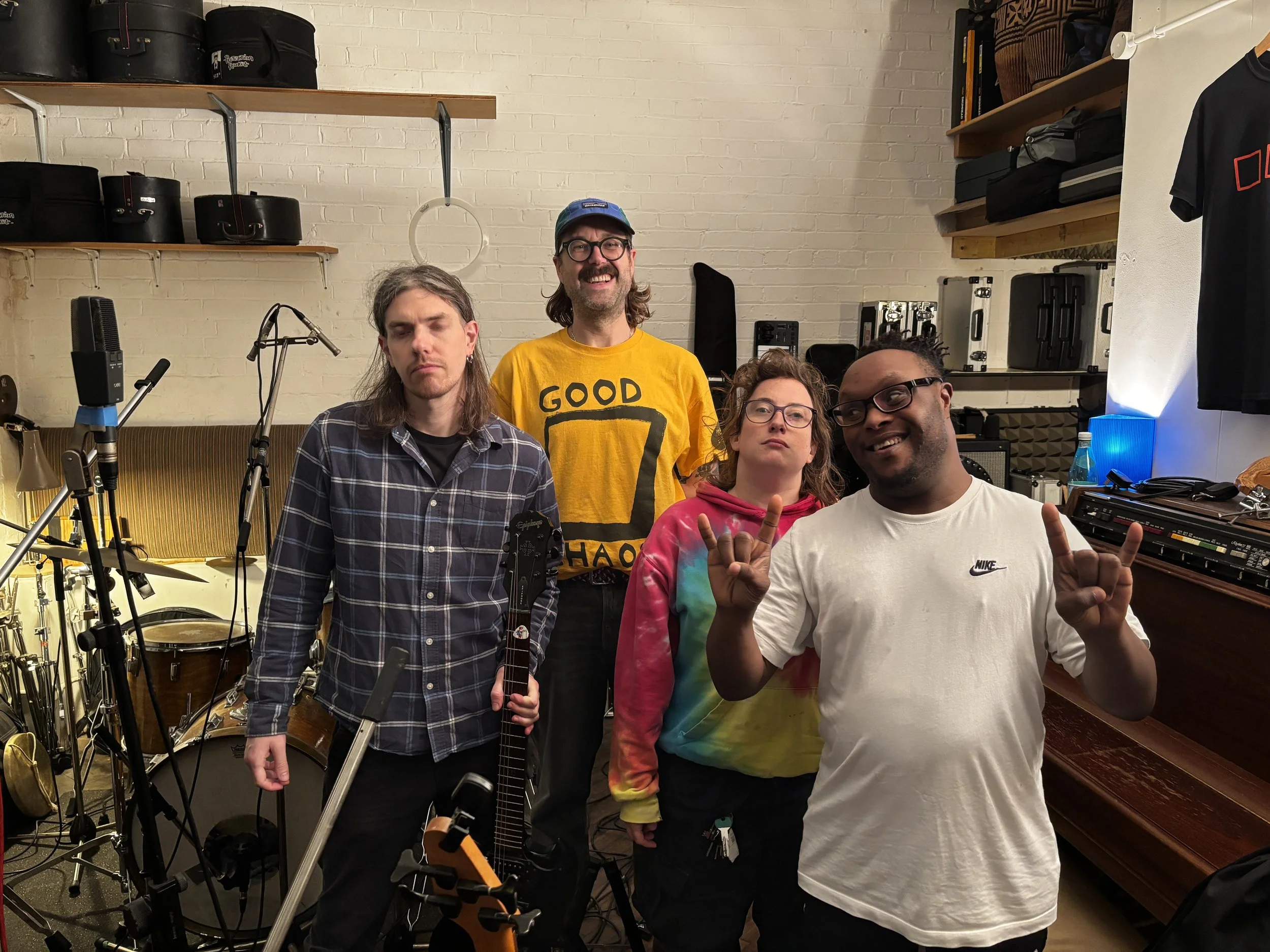 a group of people 3 male identifying people and 1 woman in a studio with microphones and drum boxes.