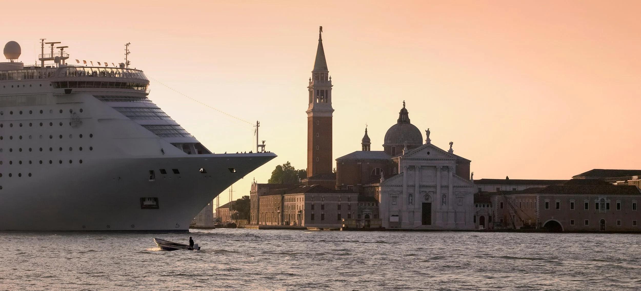 Ein großes Kreuzfahrtschiff fährt nahe an der historischen Silhouette Venedigs mit der markanten Kirche San Giorgio Maggiore vorbei. Im Vordergrund ist Wasser zu sehen.