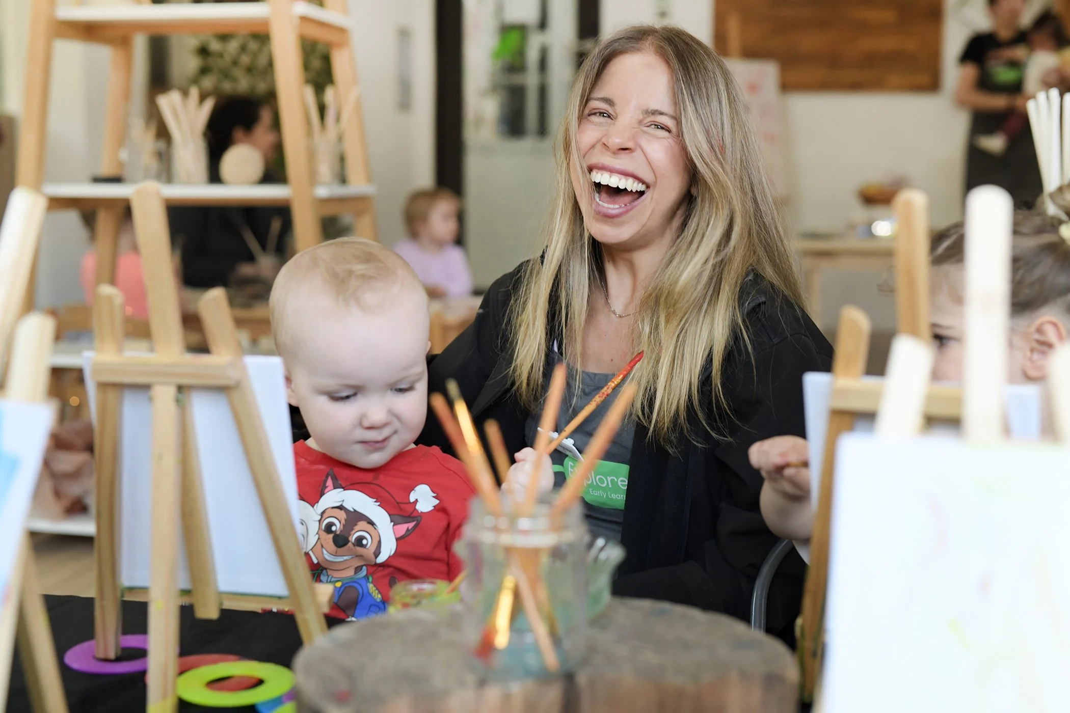 Fresh vibrant educator, laughing while helping a young child paint- paint brushes- relaxed- natural- childcare.jpg
