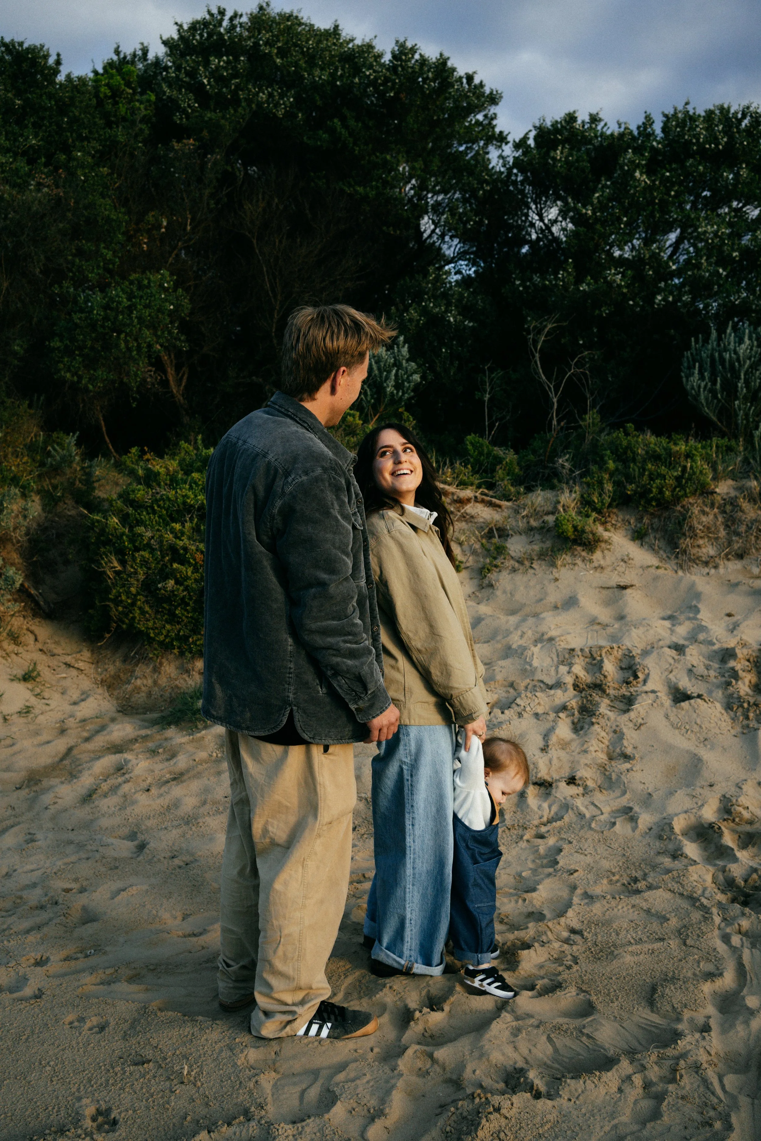 A young family stands on the sandy beach, with lush bushes and trees behind them, enjoying a moment together during sunset.